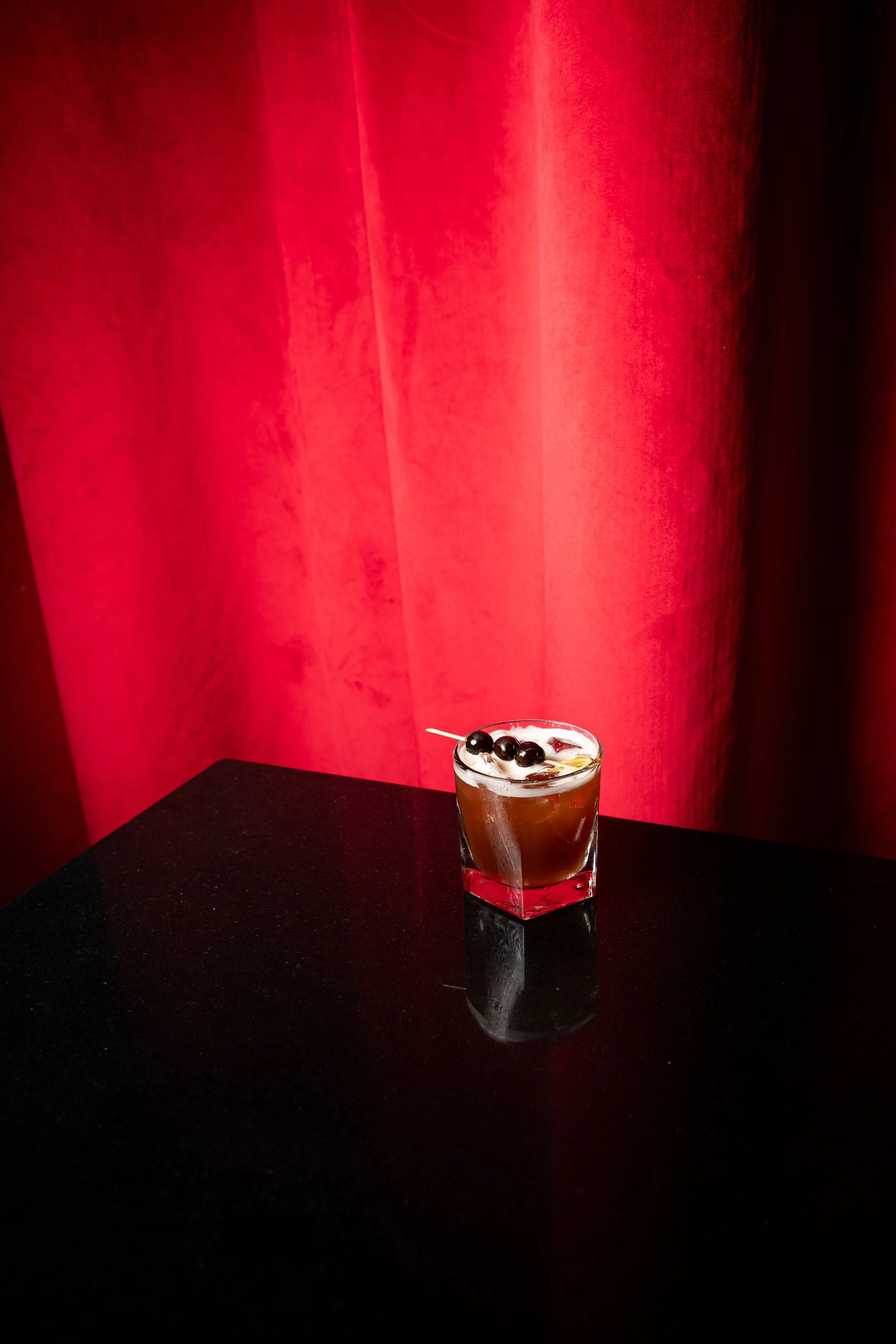 A cocktail with dark liquid, served in a clear glass with ice, garnished with black berries and a skewer, placed on a black table against a red curtain background.