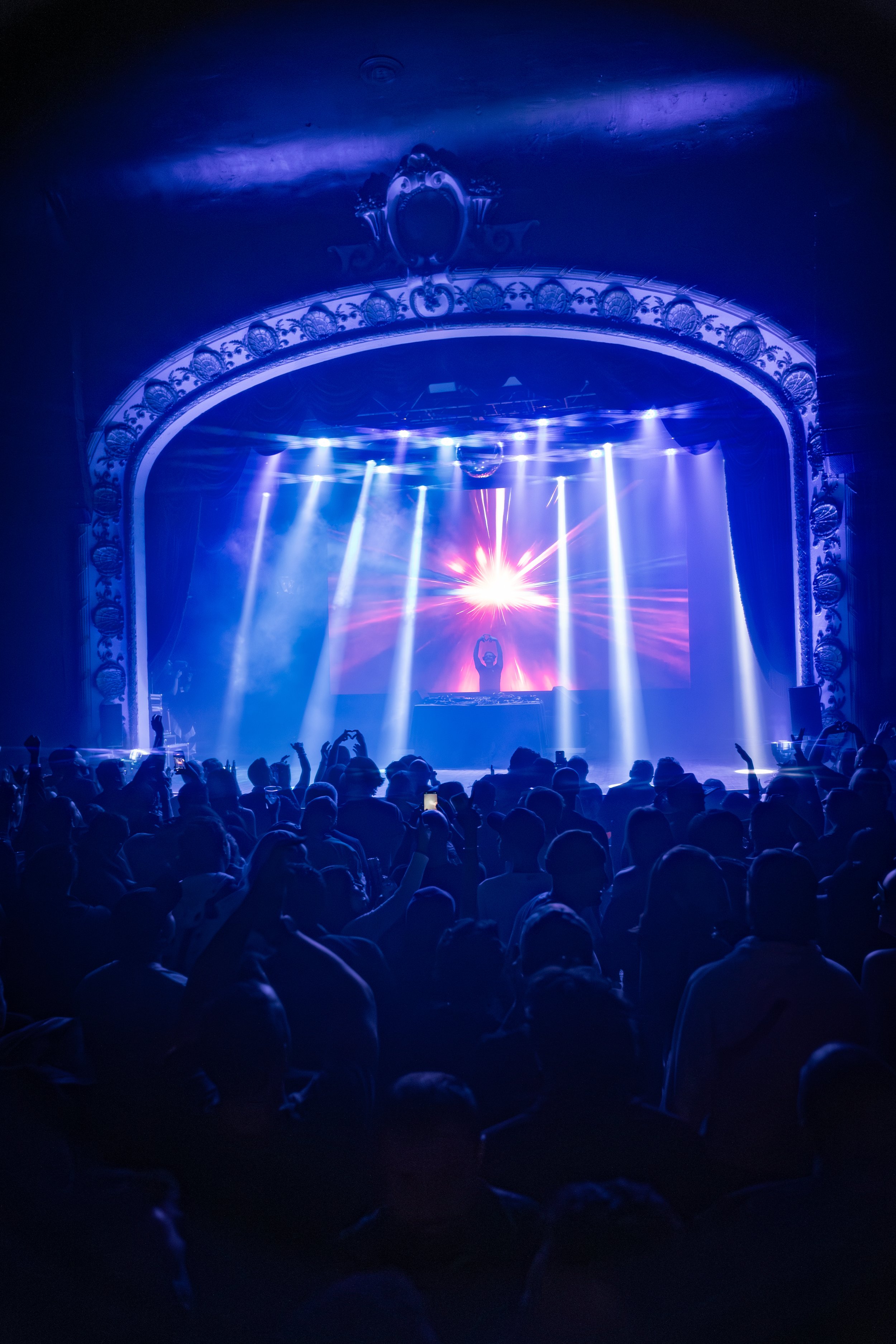 Crowd watching a DJ perform on stage with vibrant blue and purple lighting, ornate theater arch, and a bright light explosion behind the DJ.