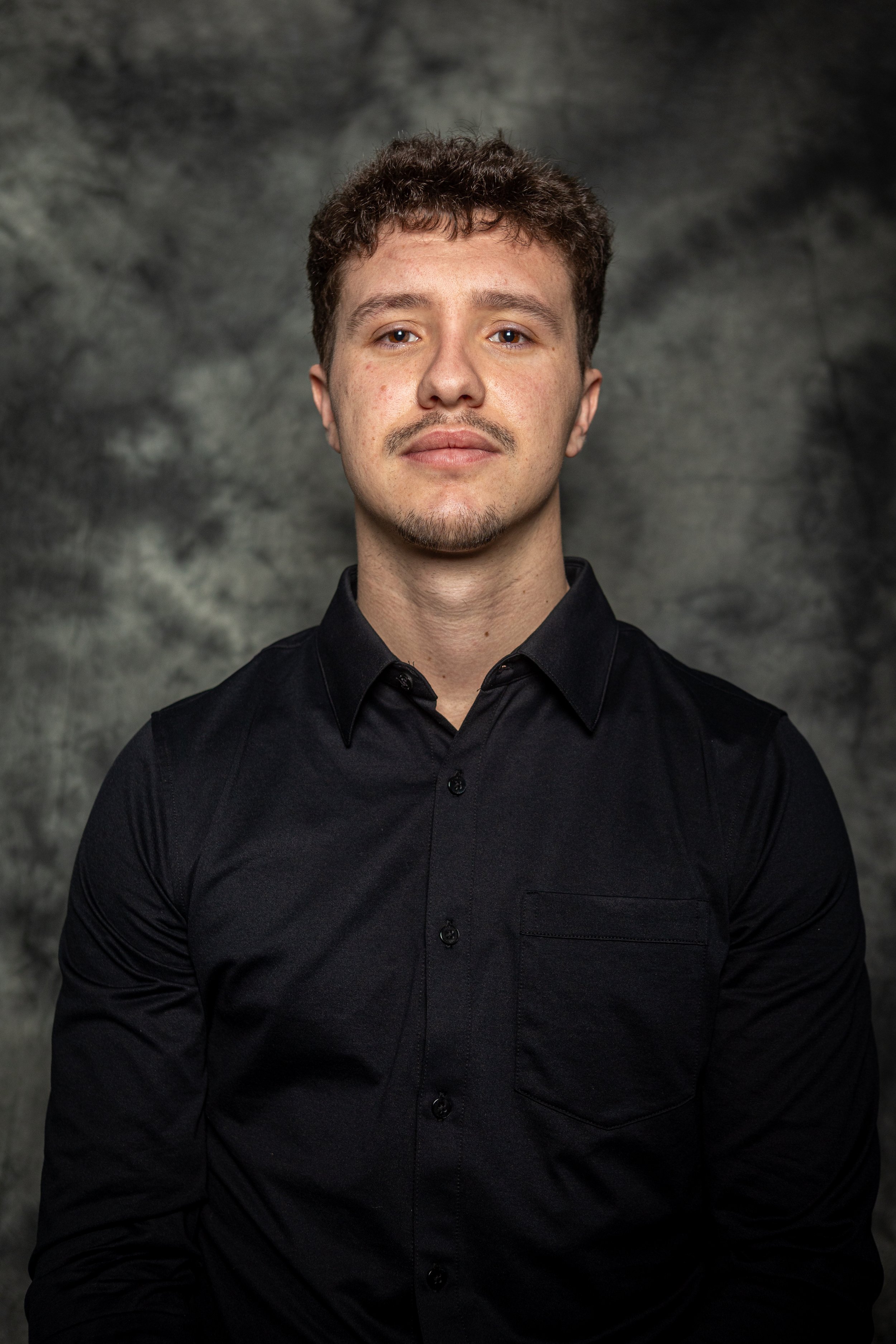 Portrait of a young man with curly brown hair and light facial hair, wearing a black button-up shirt, standing against a dark gray textured background.