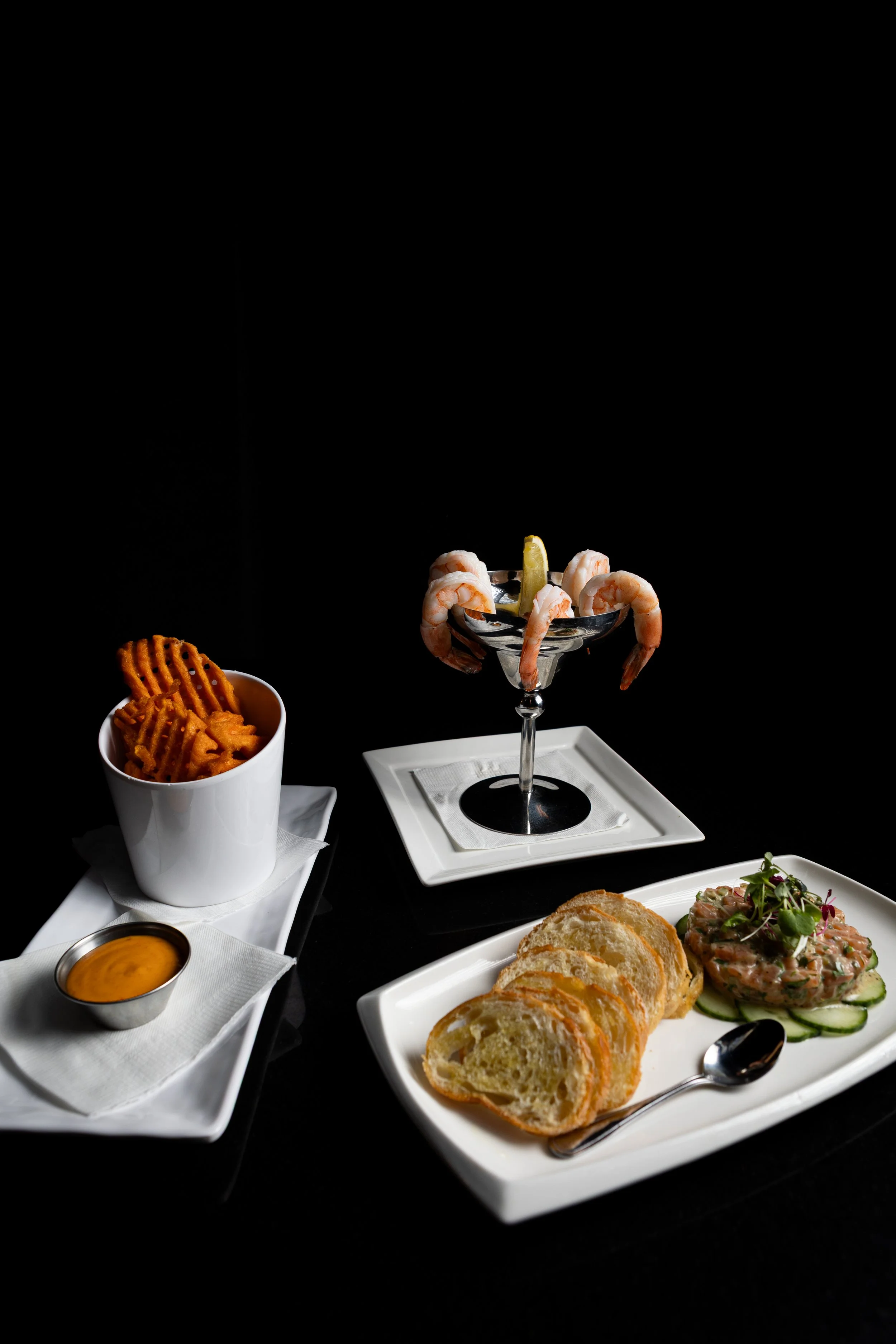 Assorted seafood appetizers including shrimp cocktail in a glass, waffle fries with dipping sauce, sliced baguette bread, and a cucumber salad with salmon and microgreens, all set against a black background.