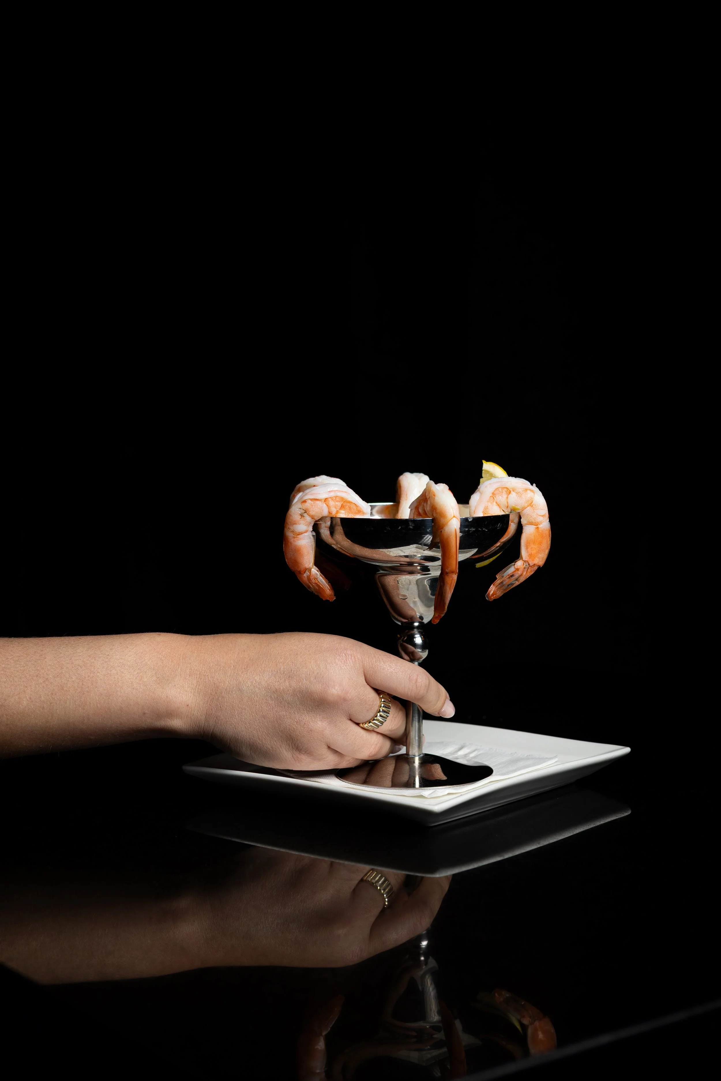 A person holding a champagne glass with shrimp draped over its rim, set on a white plate against a black background.