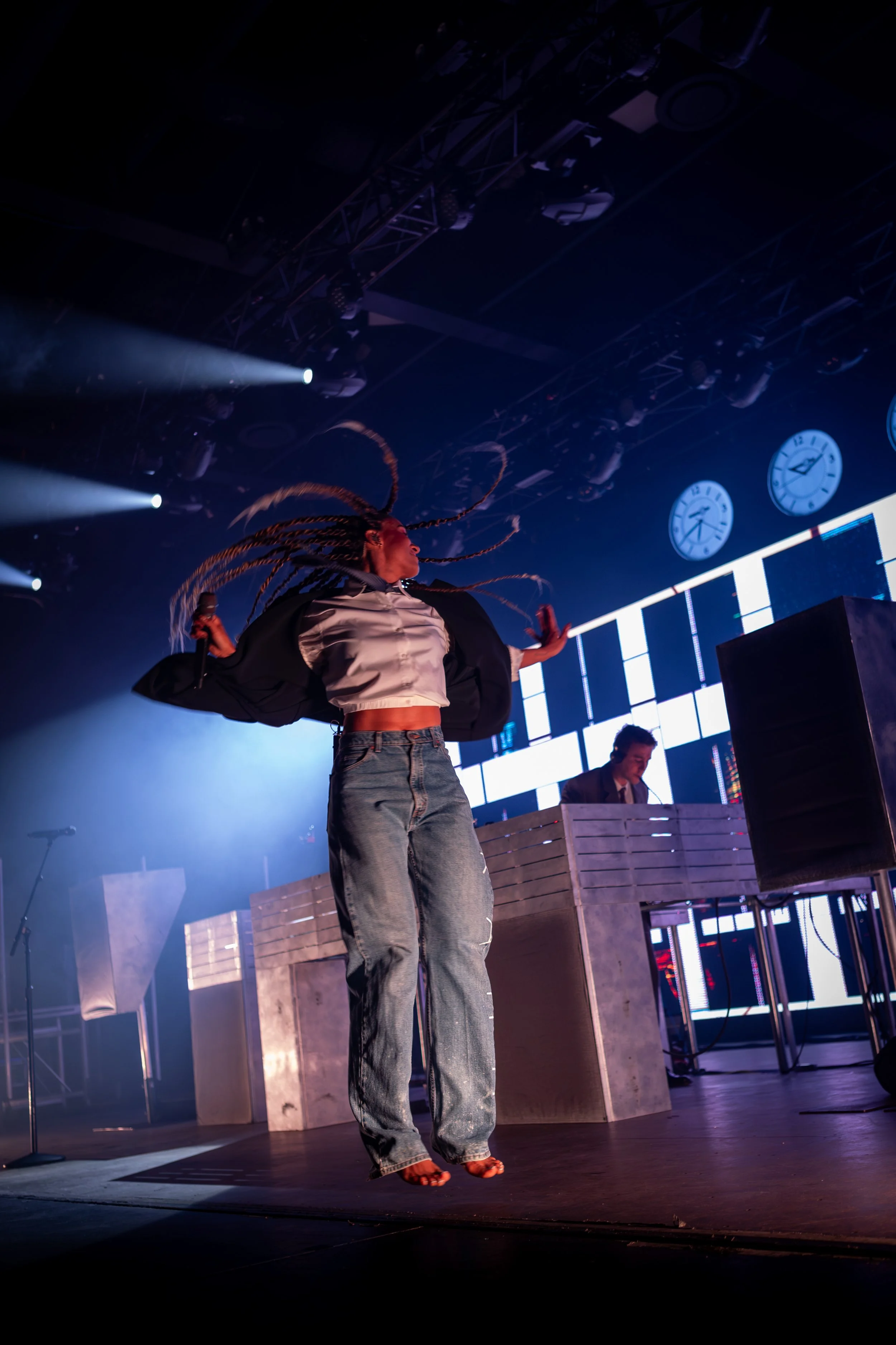 A woman dancing on stage with long braided hair, wearing a white cropped shirt, a black jacket, and jeans, with a DJ playing in the background and large clocks on the wall behind them.