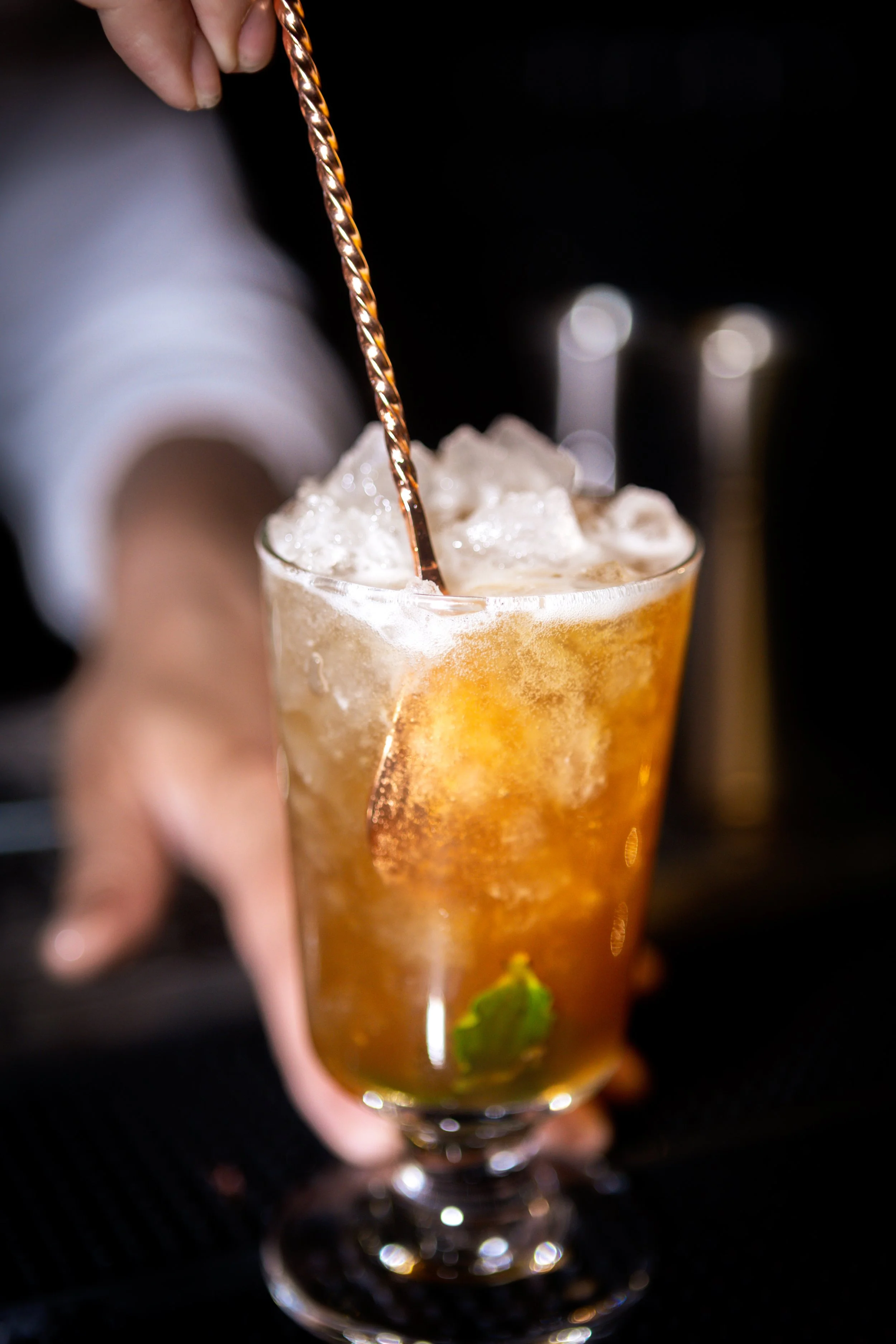 A person stirring a glass of iced tea with ice and a mint leaf at the bottom.