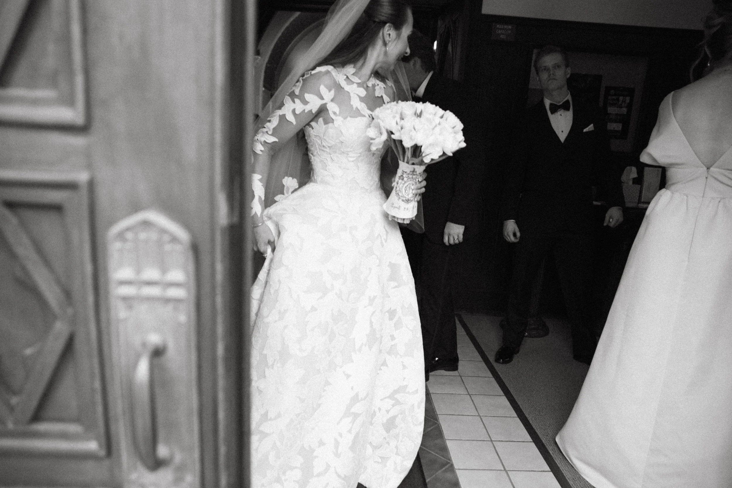 A bride in a wedding dress holding a bouquet, standing inside a building with a group of people around her, including someone in a tuxedo and others in formal dresses.