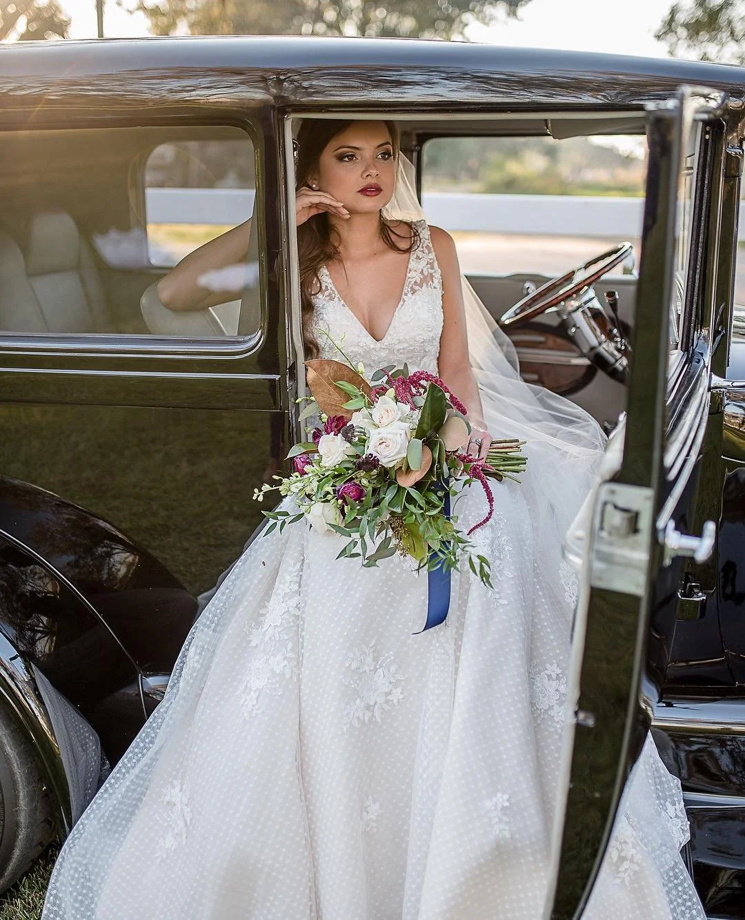 Bride sitting in vintage car, holding a bouquet of flowers, wearing a white wedding dress with lace details, with a contemplative expression.