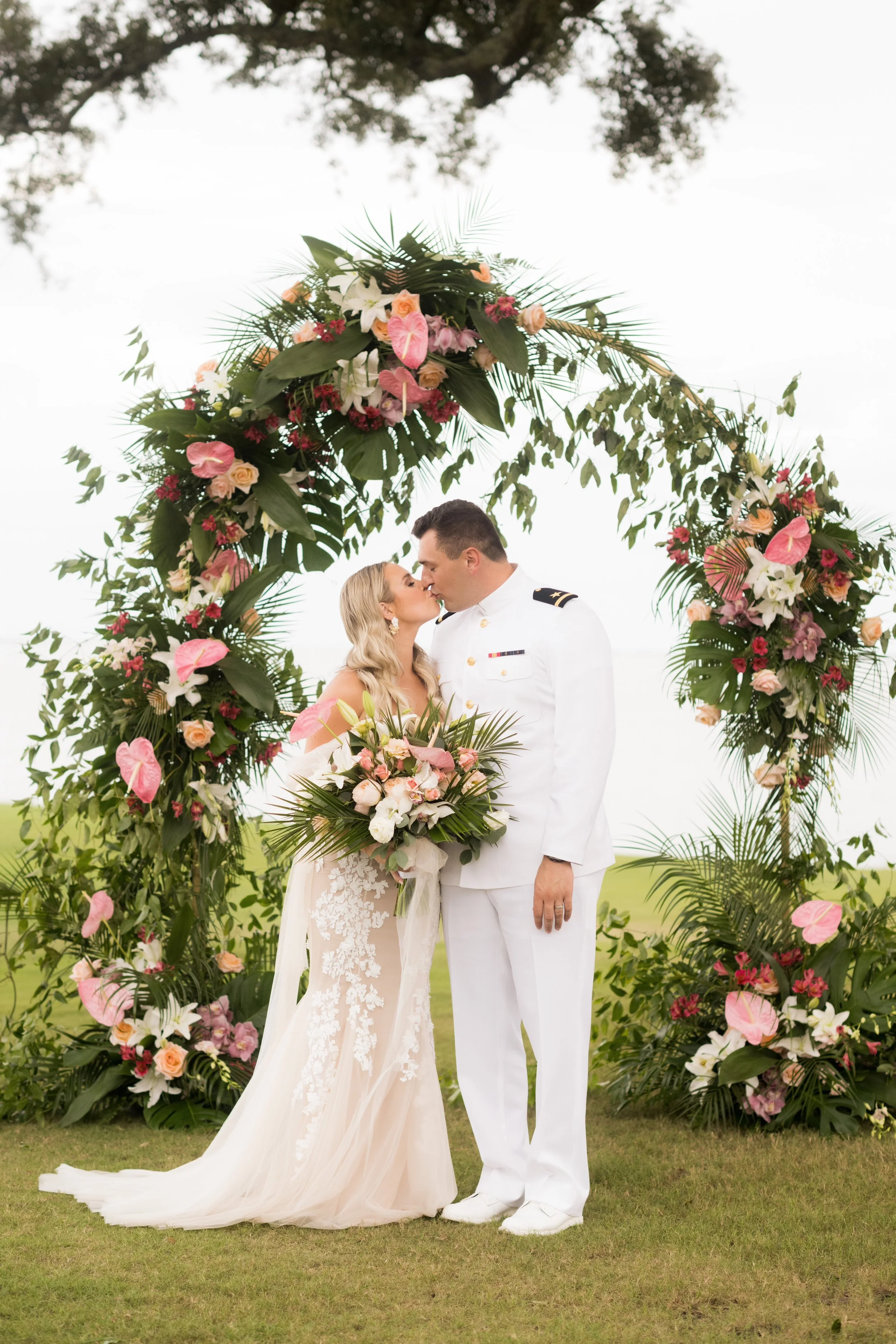 A bride and groom share a kiss under a floral arch on their wedding day outdoors.