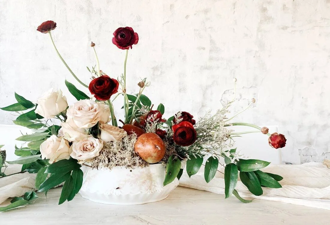 A floral arrangement with cream and red roses, ranunculus, greenery, moss, a pomegranate, and other flowers in a white ceramic bowl on a wooden table against a white brick wall.