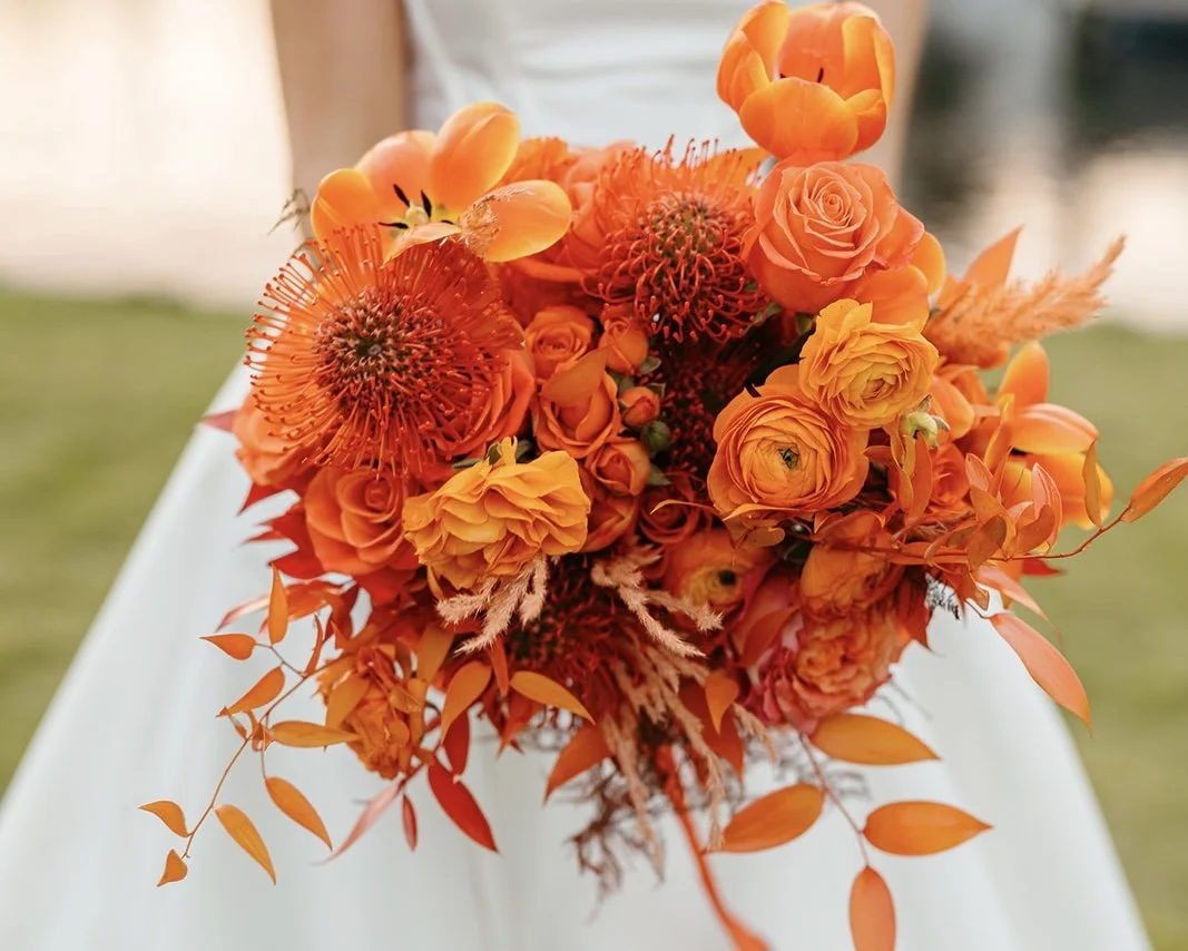 Orange flower bouquet with roses, lilies, and greenery held by a person in a white dress.