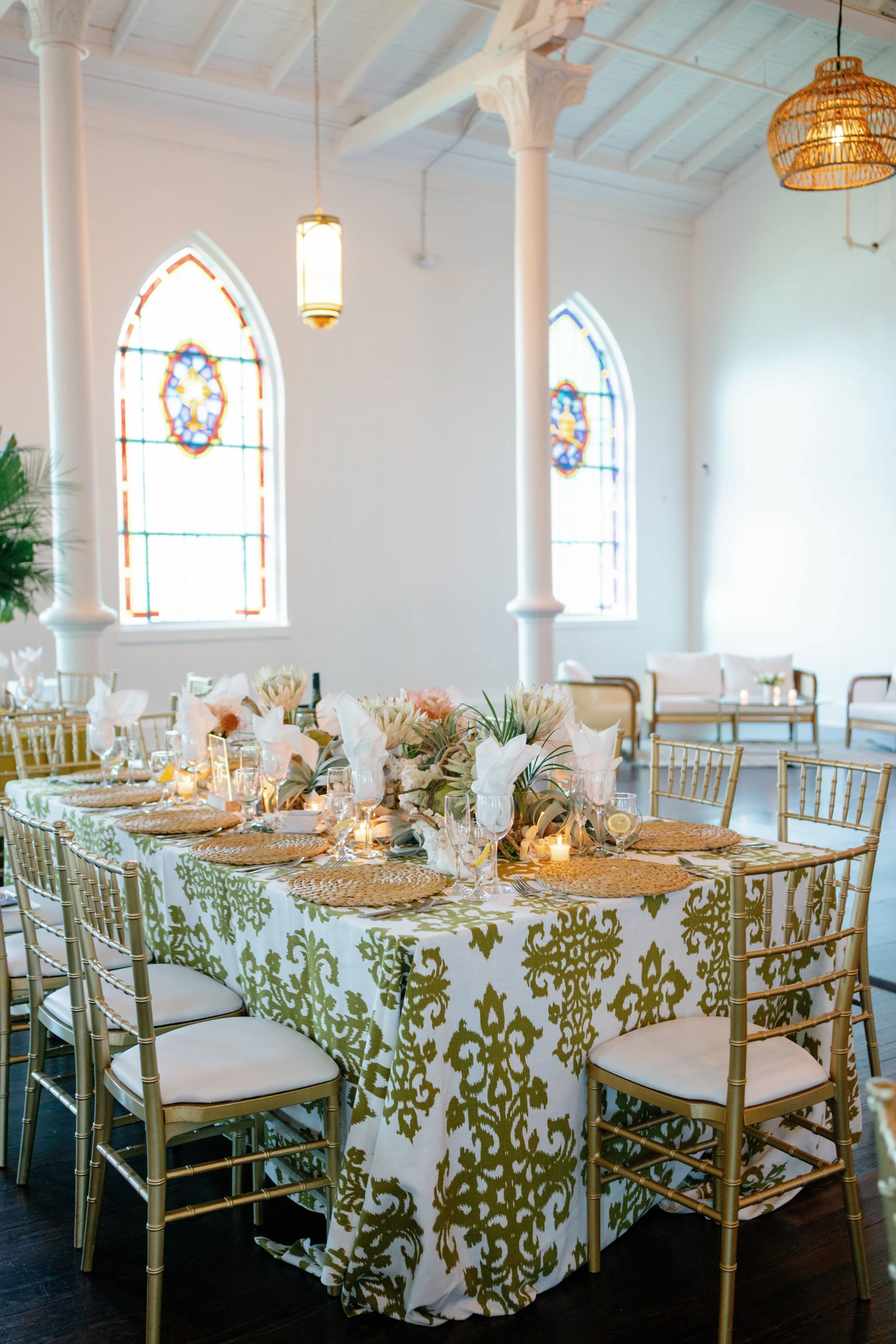 Wedding reception table with gold chairs, white and gold tablecloth, floral centerpiece, candles, and elegant place settings inside a decorated church with stained glass windows.
