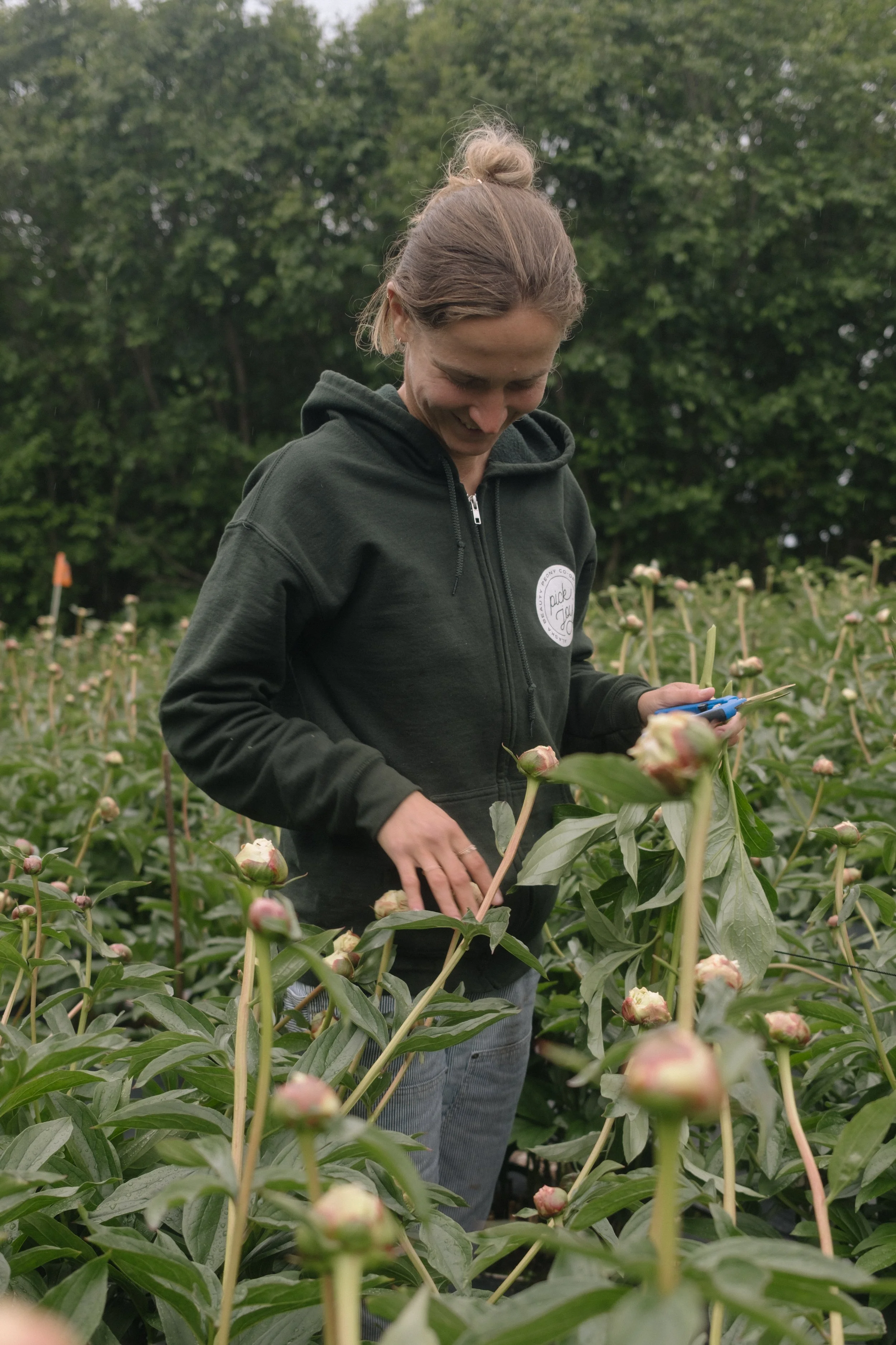 A woman in a black hoodie and jeans looks down and smiles while examining or working on peony flowers in a lush green outdoor garden or field.