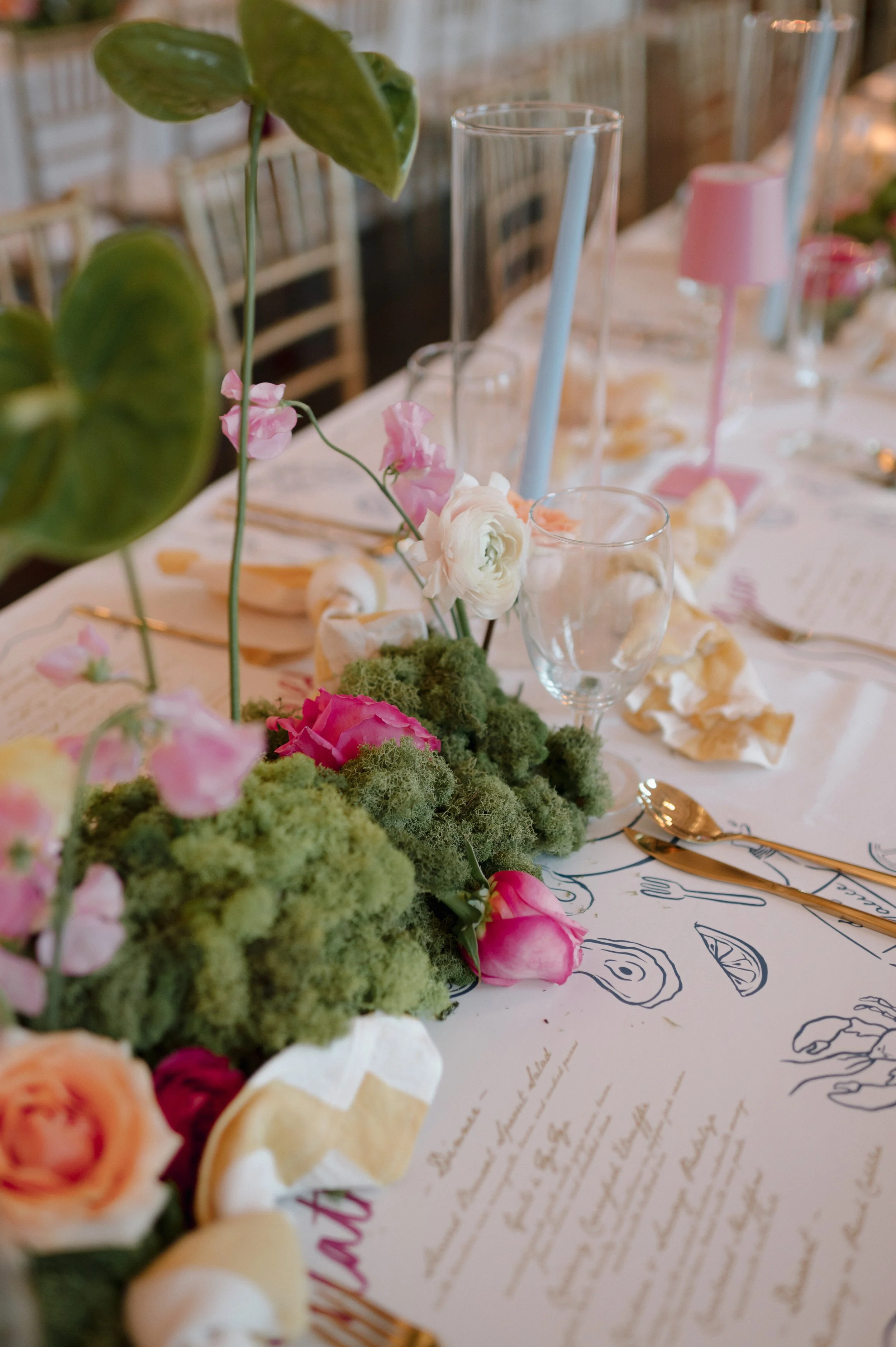 A table decorated with pink and white flowers, greenery, wine glasses, gold utensils, and pink candles in a bright, elegant setting.