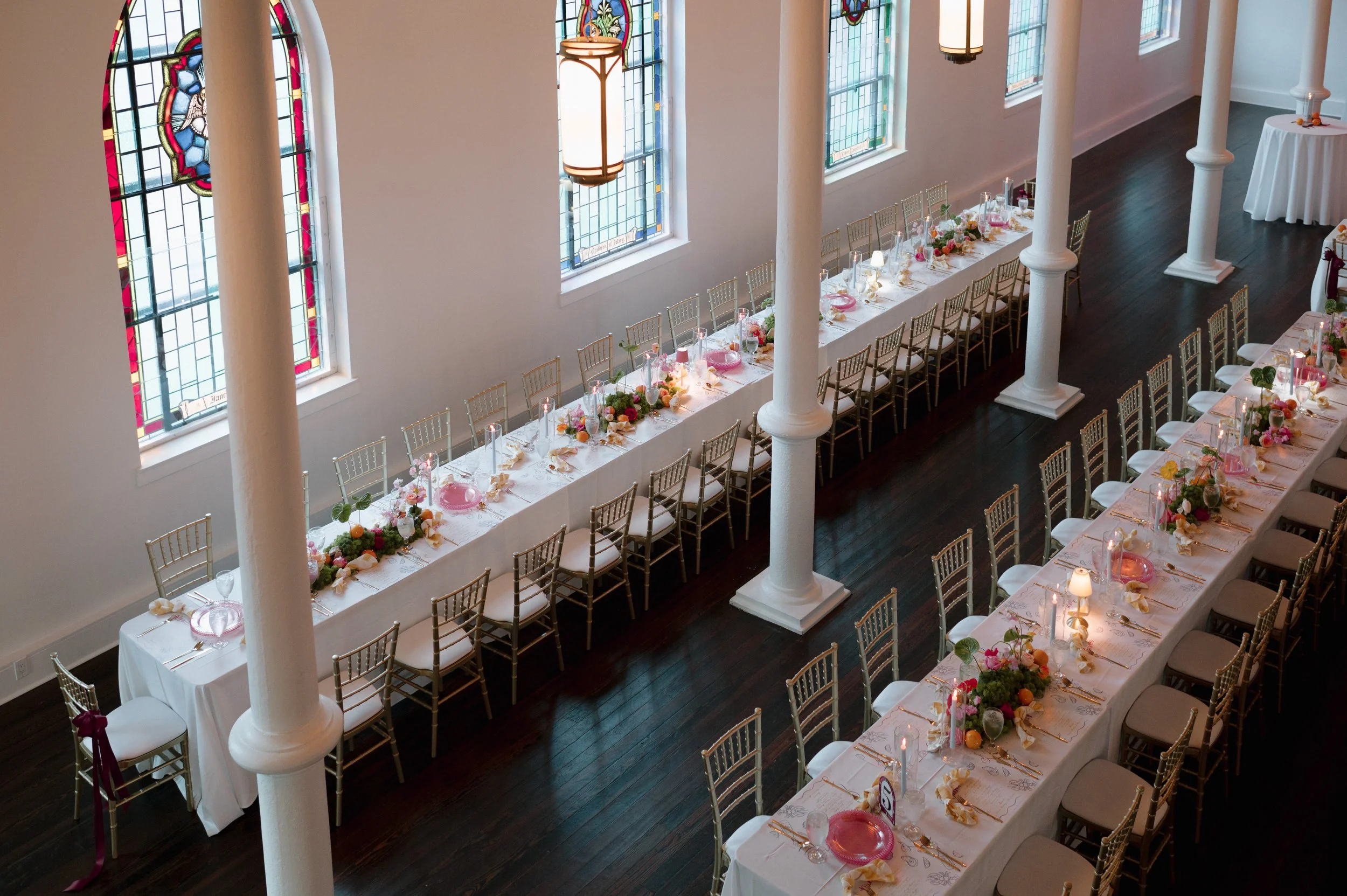 Elegant wedding reception setup with long tables decorated with floral centerpieces, candles, and pastel-colored decorations inside a church with stained glass windows and white columns.