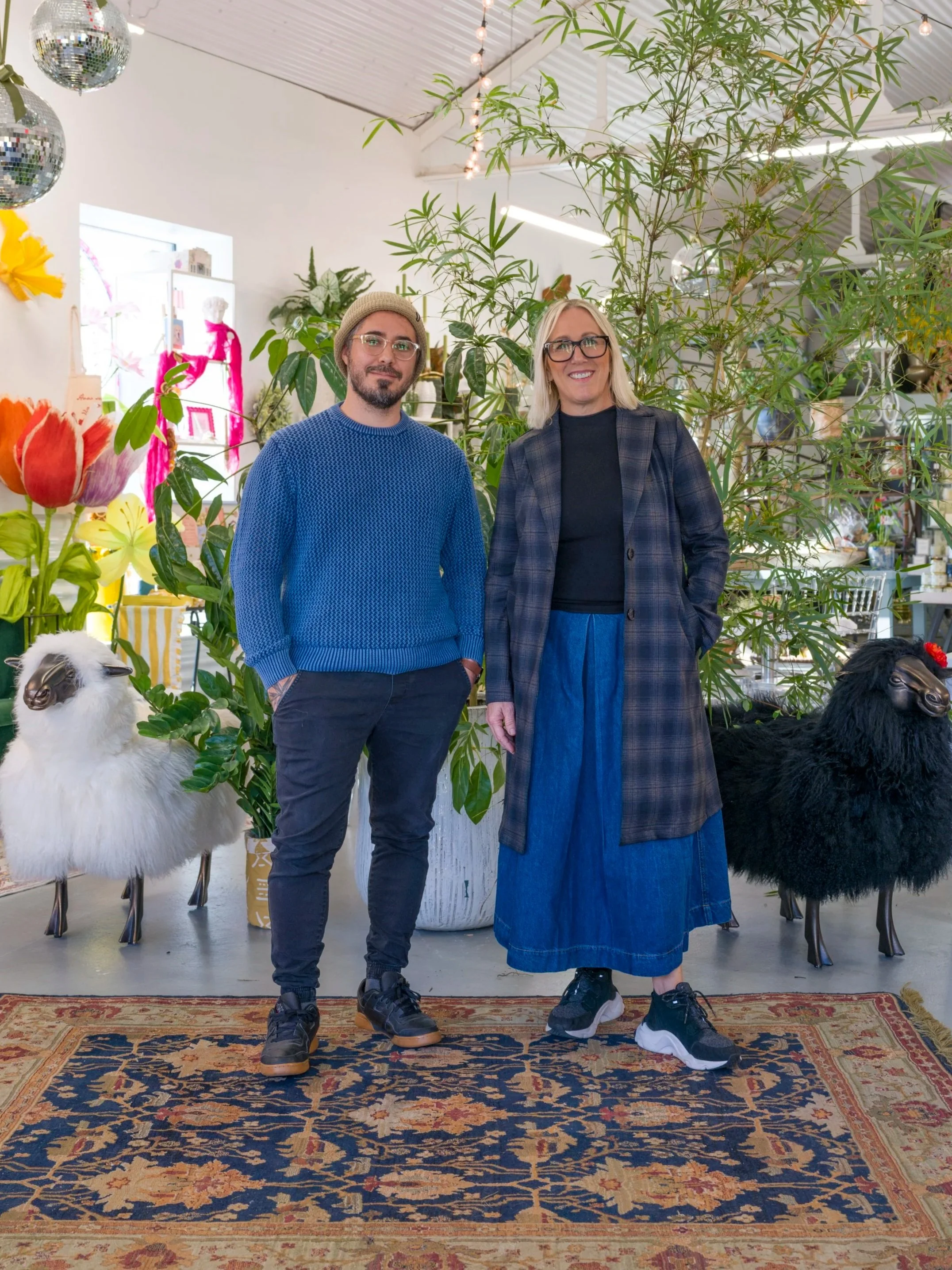 Two people, a man and a woman, standing indoors in front of green plants and decorative sheep. The man is wearing a blue sweater, dark pants, and a beige beanie. The woman is dressed in a plaid coat, black top, denim skirt, and sneakers. The room is decorated with flowers, a Persian rug, and shiny disco balls.