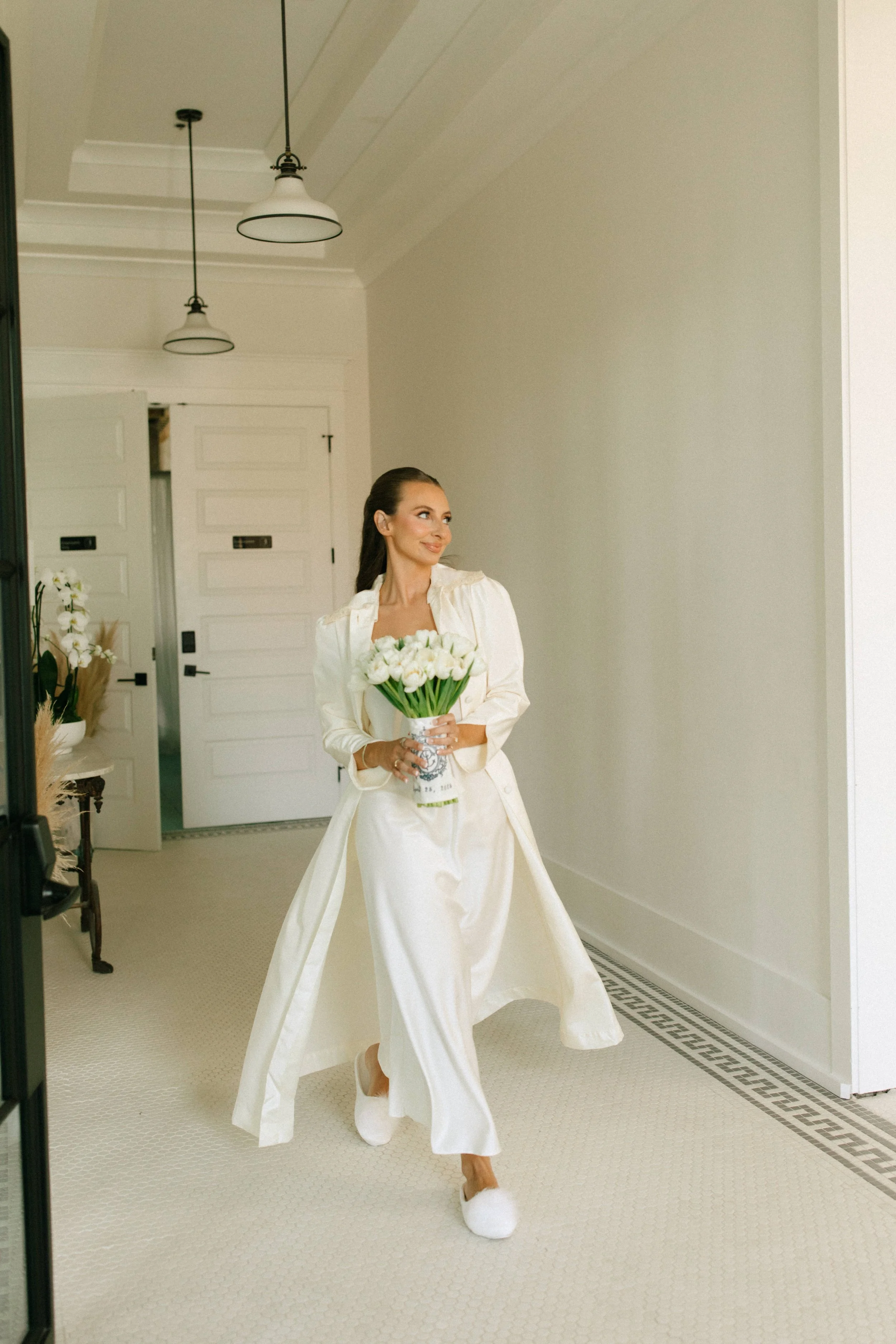 A woman dressed in a white outfit holding a bouquet of white flowers, walking indoors in a well-lit room with white walls and ceiling, with modern light fixtures hanging from the ceiling.