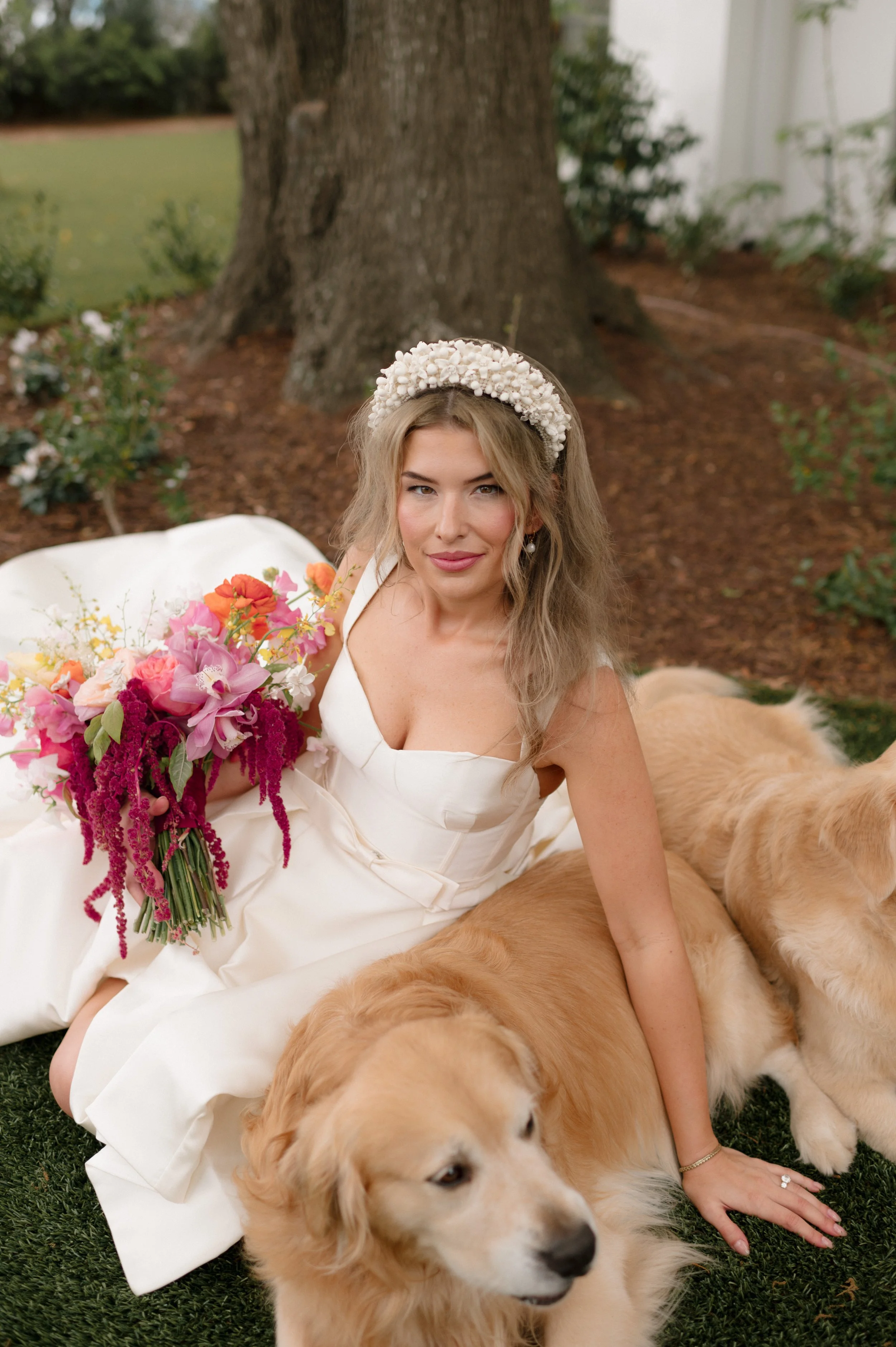 A woman in a white dress with a floral headpiece sitting on grass, holding a colorful bouquet of flowers, with two golden retrievers nearby, in a garden setting.