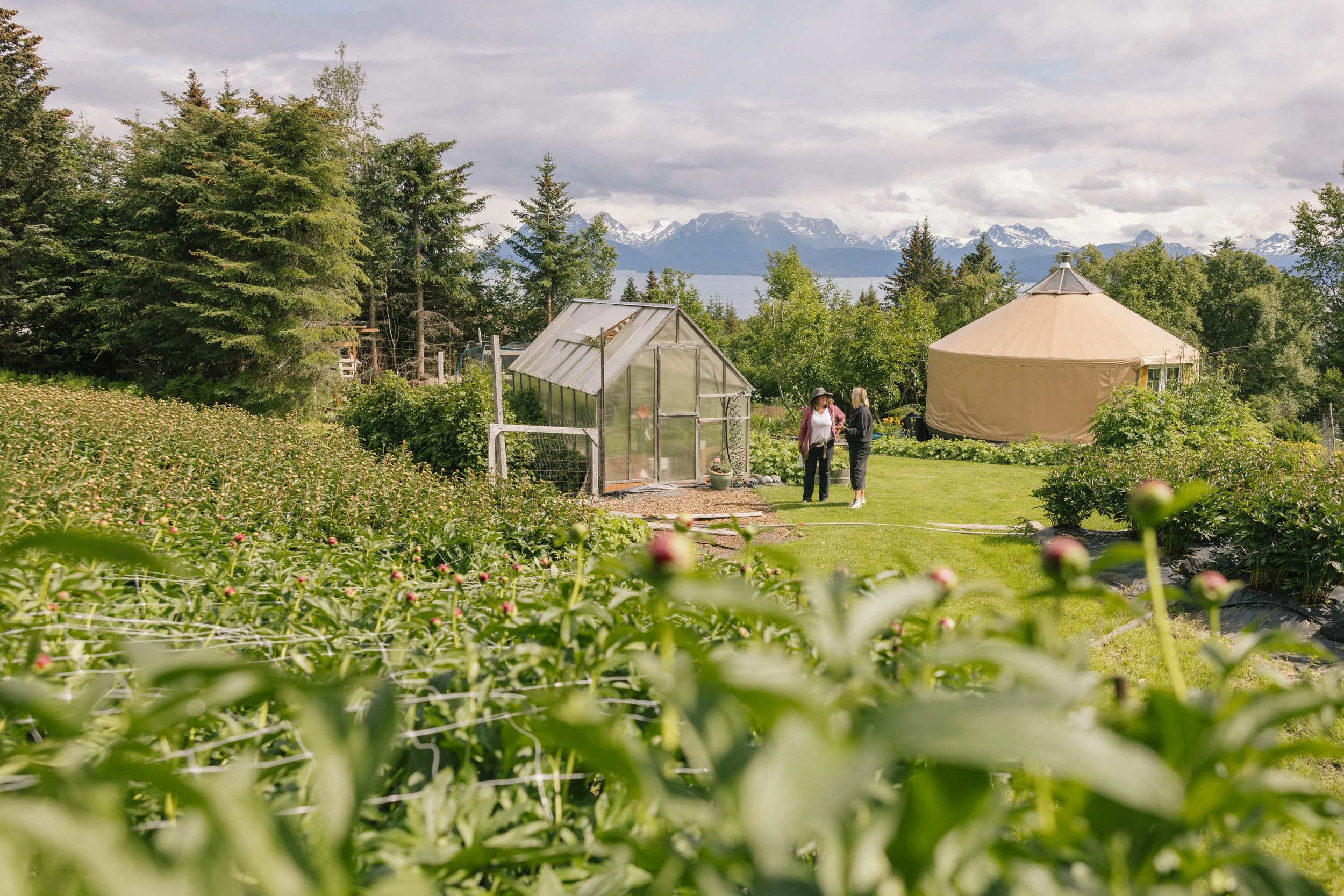 Two women stand and converse near a greenhouse and a yurt in a lush garden with mountains in the background.