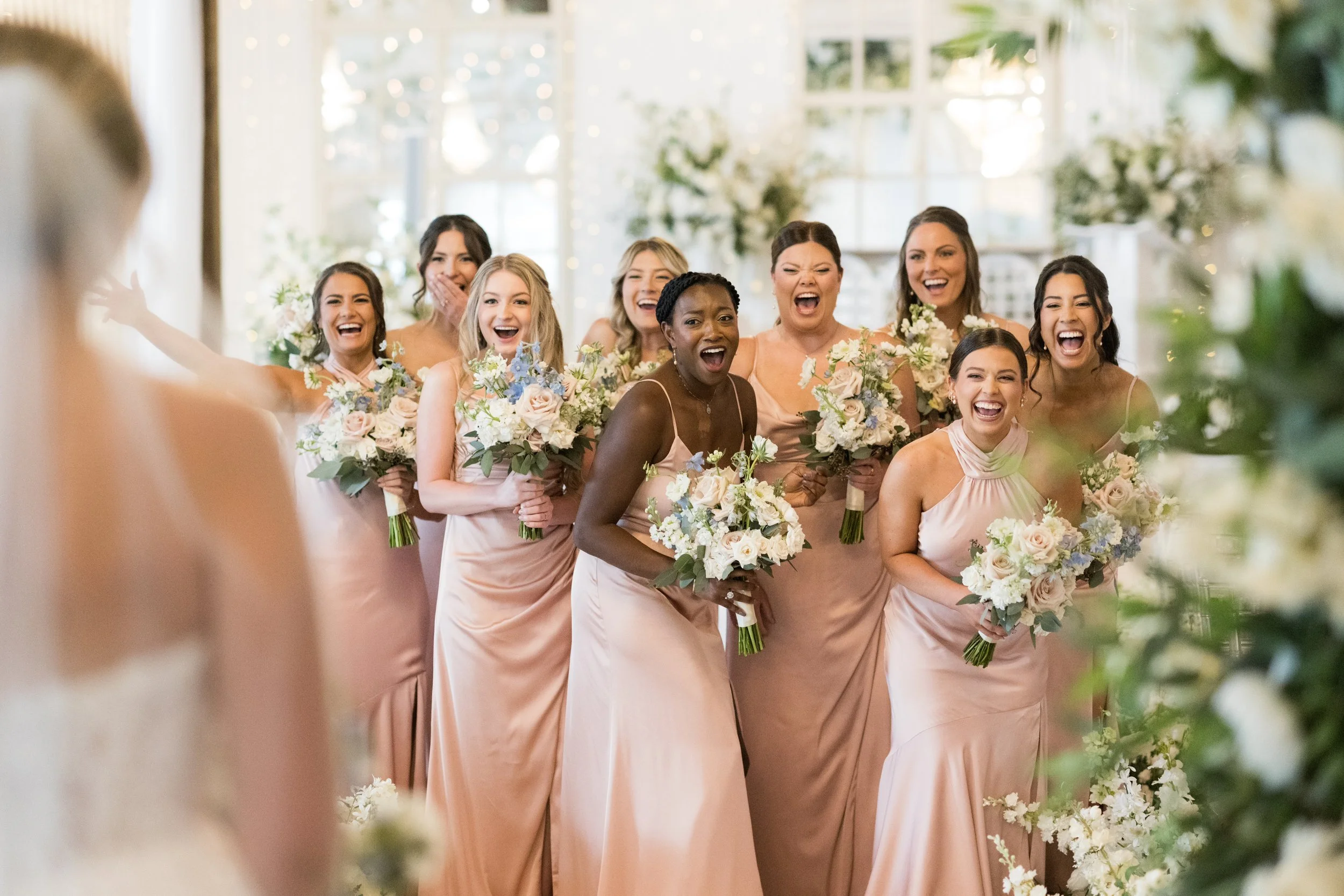 A group of women in matching peach-colored dresses holding bouquets of flowers, laughing and smiling together at a wedding.