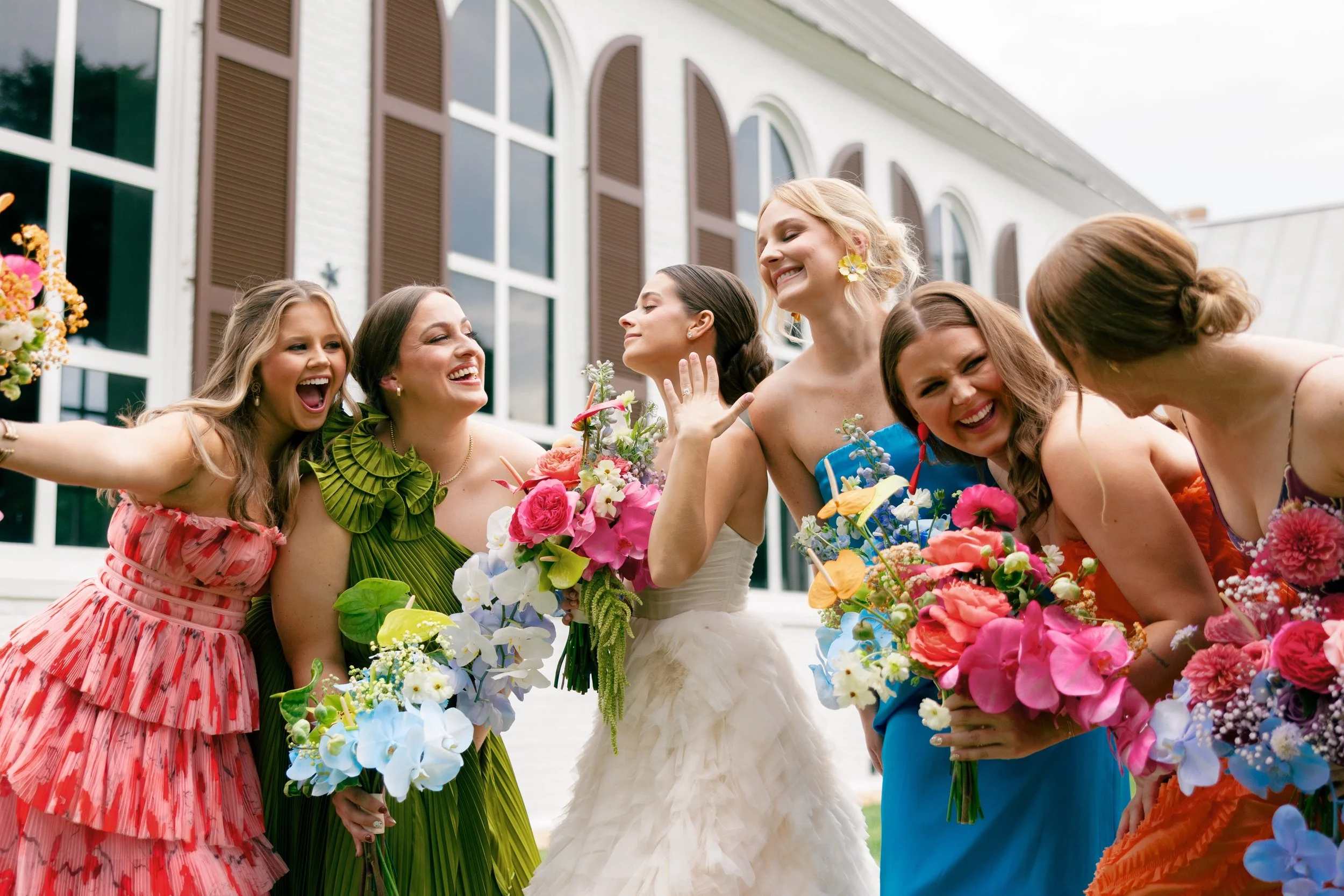 A group of women in colorful dresses holding flowers, smiling and celebrating outdoors near a white building with large windows.