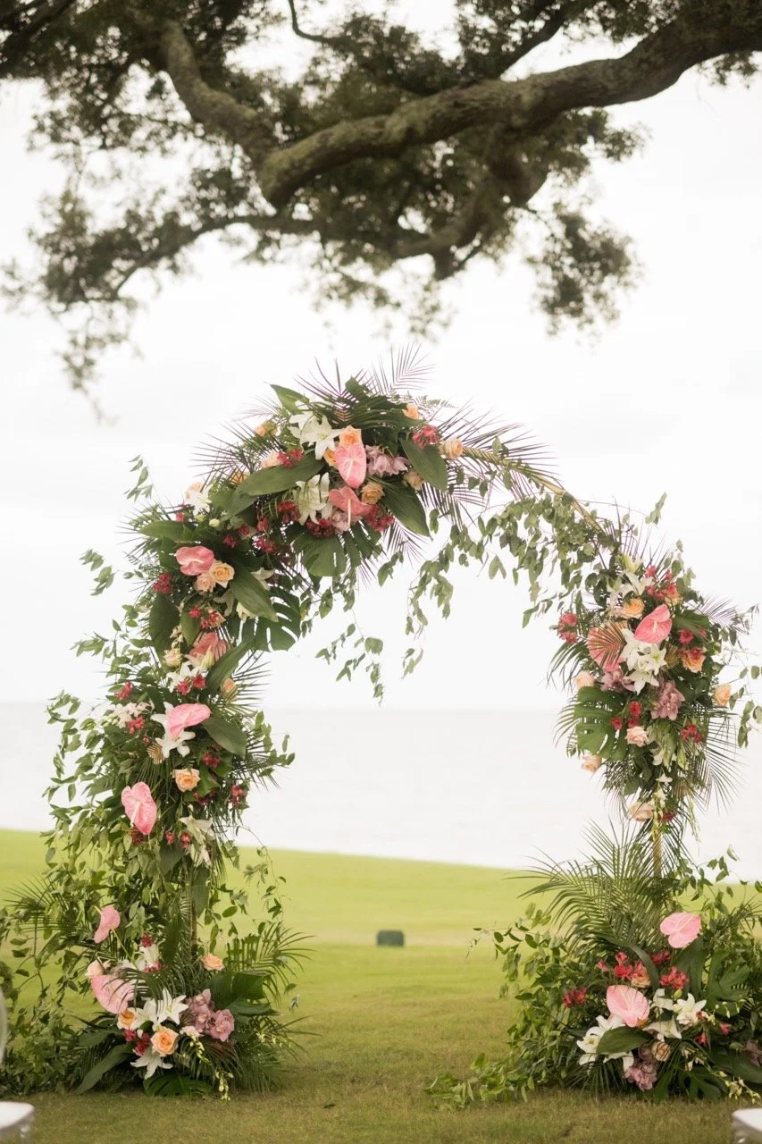 A floral wedding arch decorated with pink, white, and peach flowers, and greenery, set outdoors with a grassy area and tree overhead.