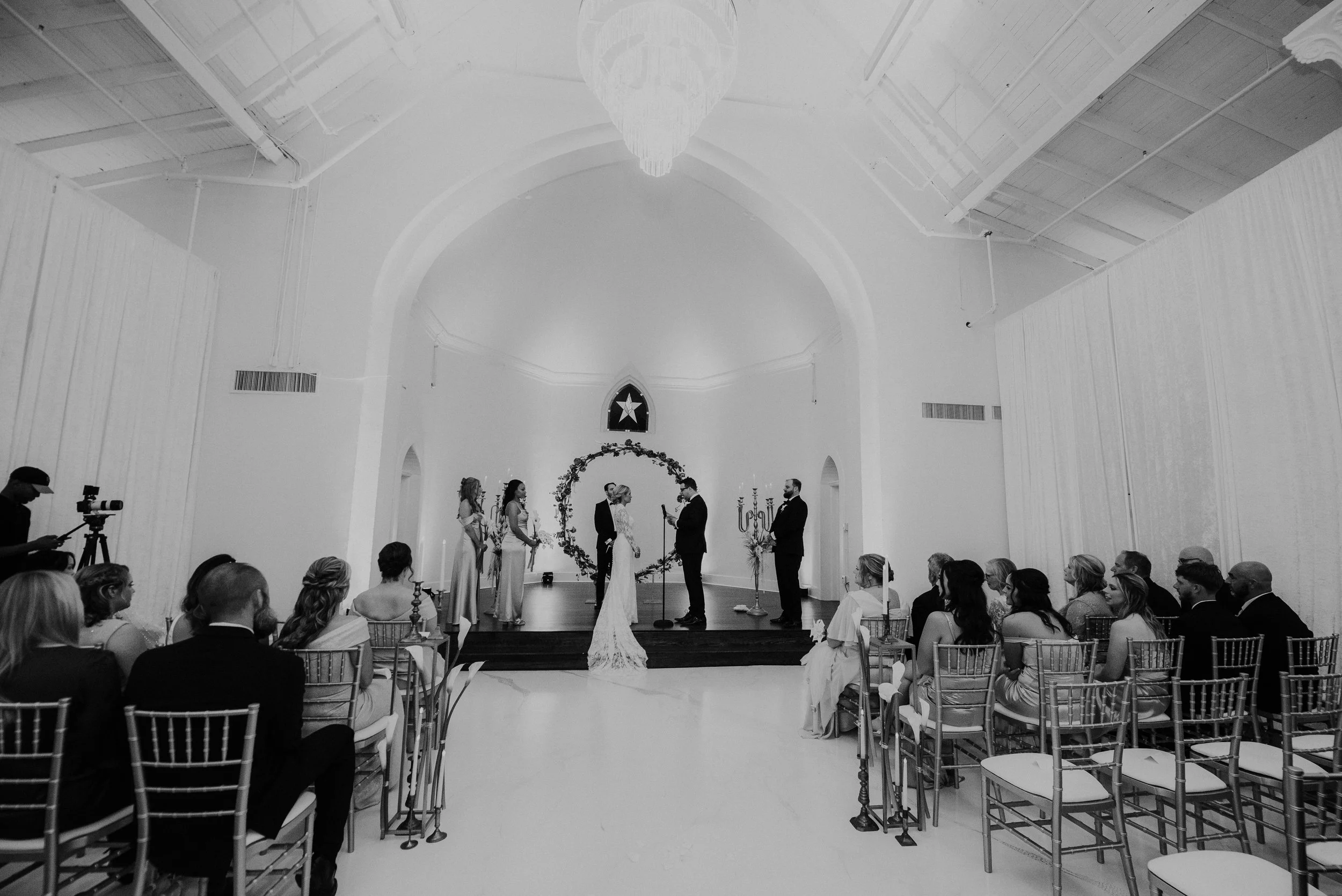 A wedding ceremony inside a white chapel with an arched ceiling, officiant and bride and groom at the altar, surrounded by bridesmaids and groomsmen, guests seated on chairs, and a photographer recording the event.