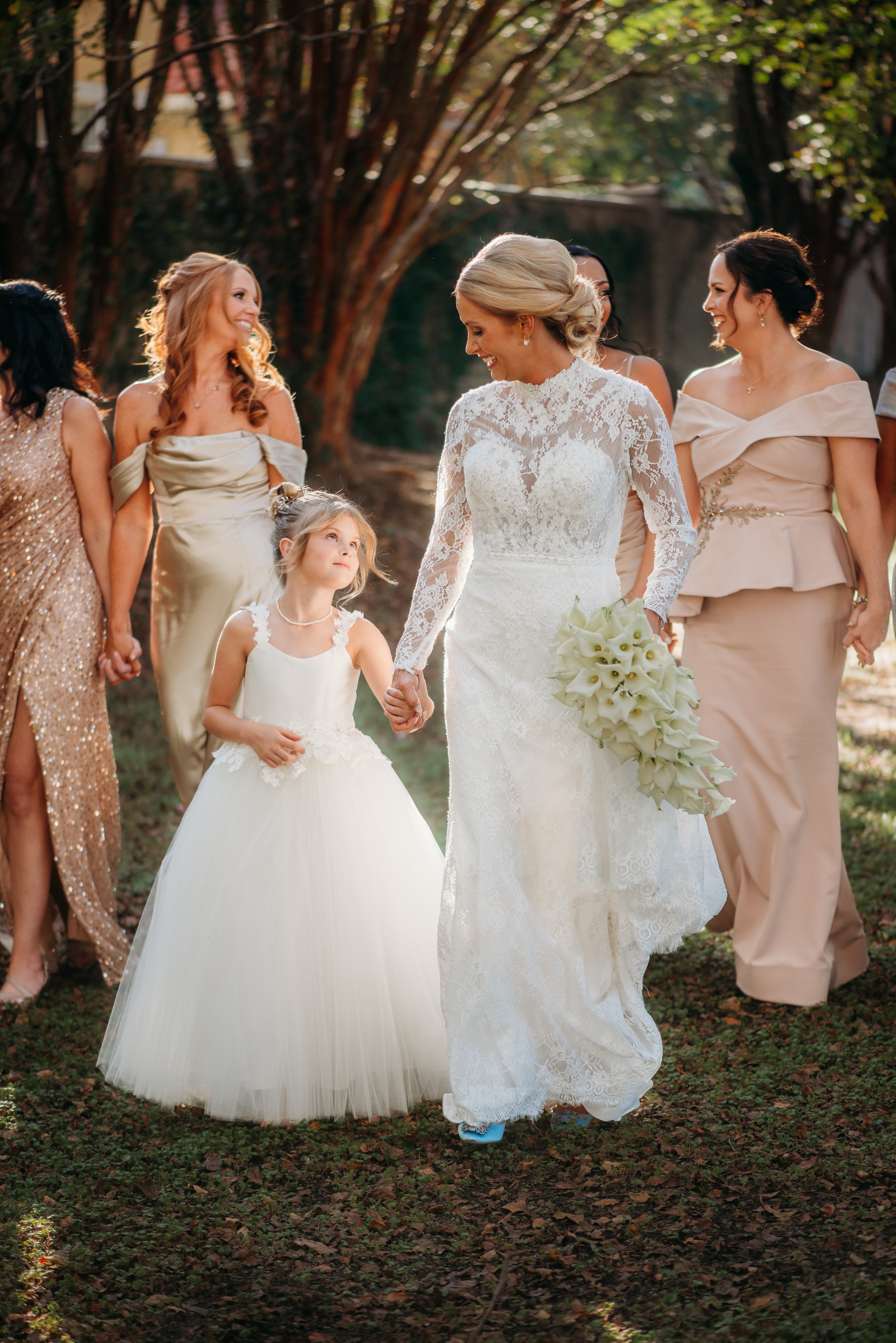 A bride in a white lace wedding dress holding a bouquet, walking hand-in-hand with a young girl in a white dress, surrounded by smiling women in elegant dresses outdoors during sunset.