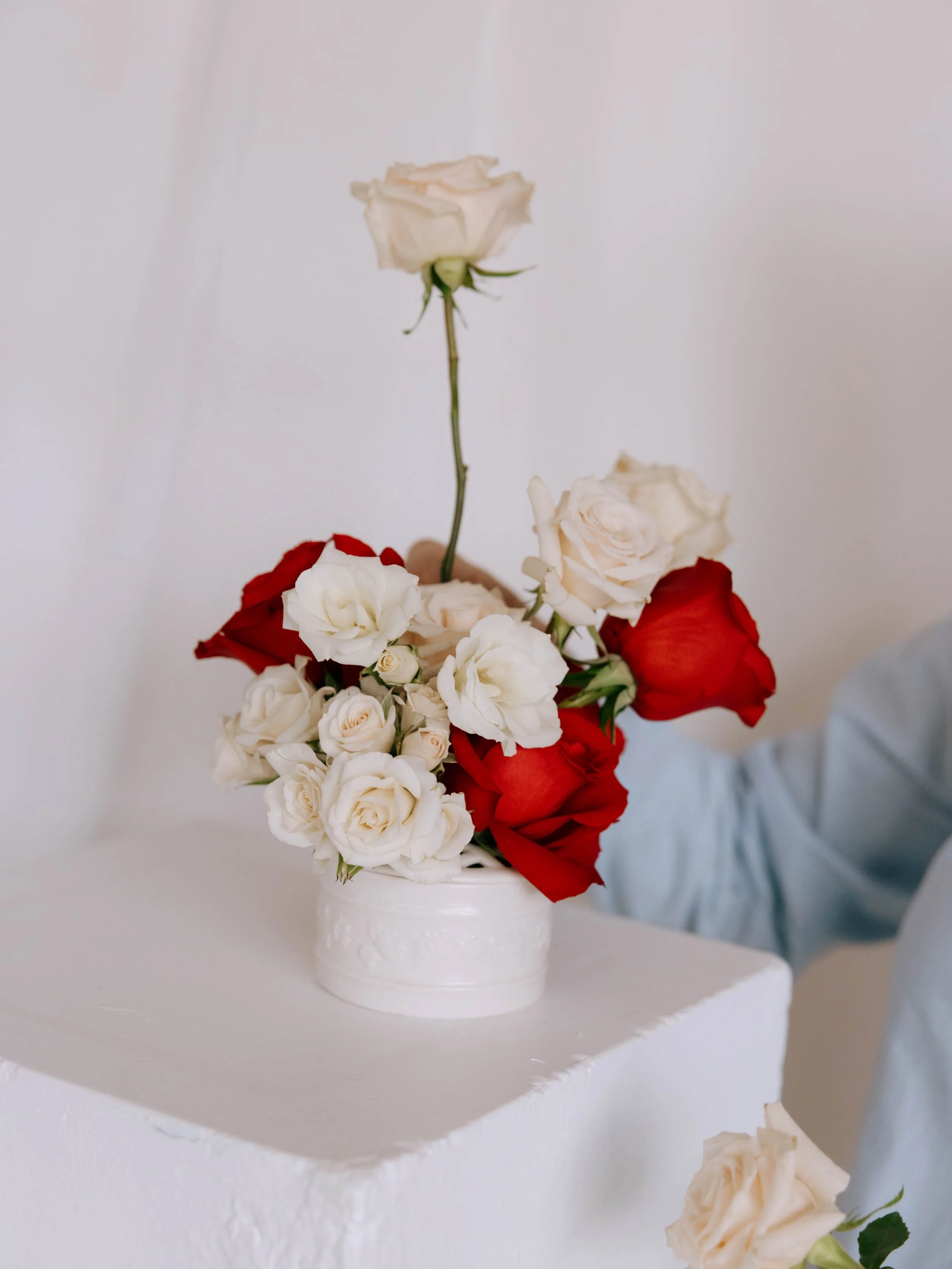 A white vase with intricate patterns containing a bouquet of red, white, and cream roses, placed on a white pedestal.
