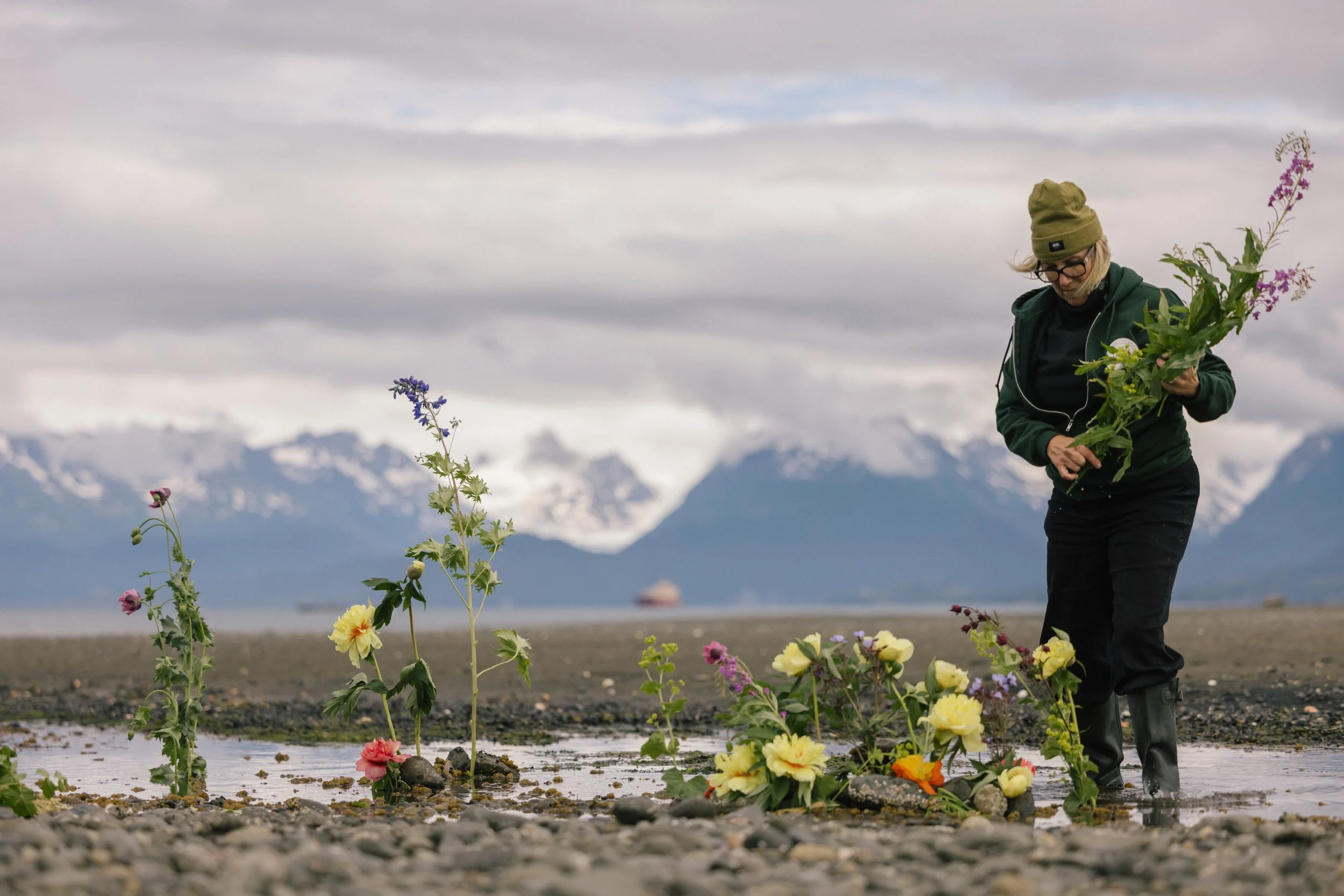 A woman in outdoor gear, including a green hoodie, khaki beanie, and rubber boots, arranging flowers on a rocky shoreline with snow-capped mountains in the background.