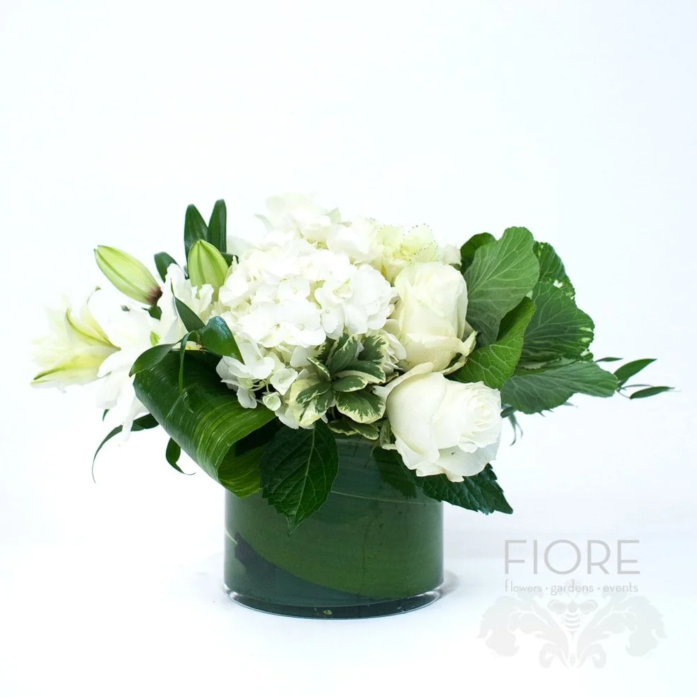 A floral arrangement of white flowers and green leaves in a dark green glass vase.