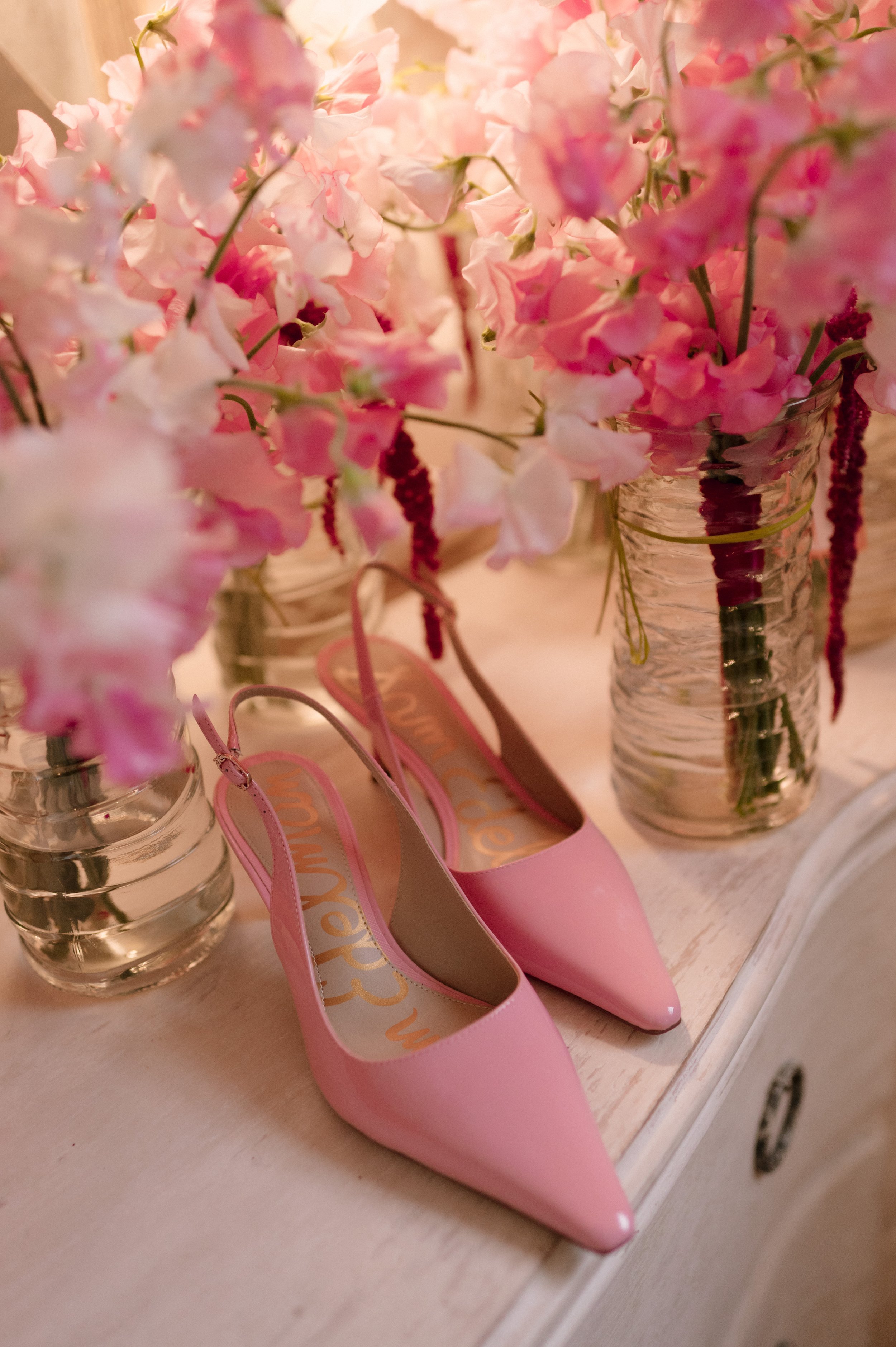 Pink high-heeled shoes next to glass vases filled with pink flowers on a white table.