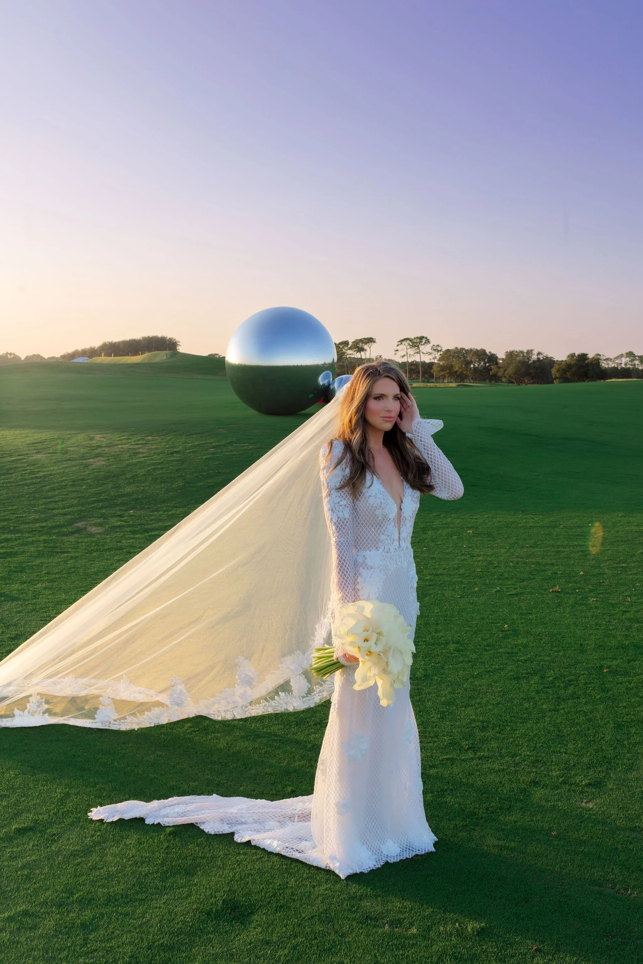 A woman in a white wedding gown holding a bouquet of white flowers stands on a green field at sunset, with large reflective spherical sculptures in the background.