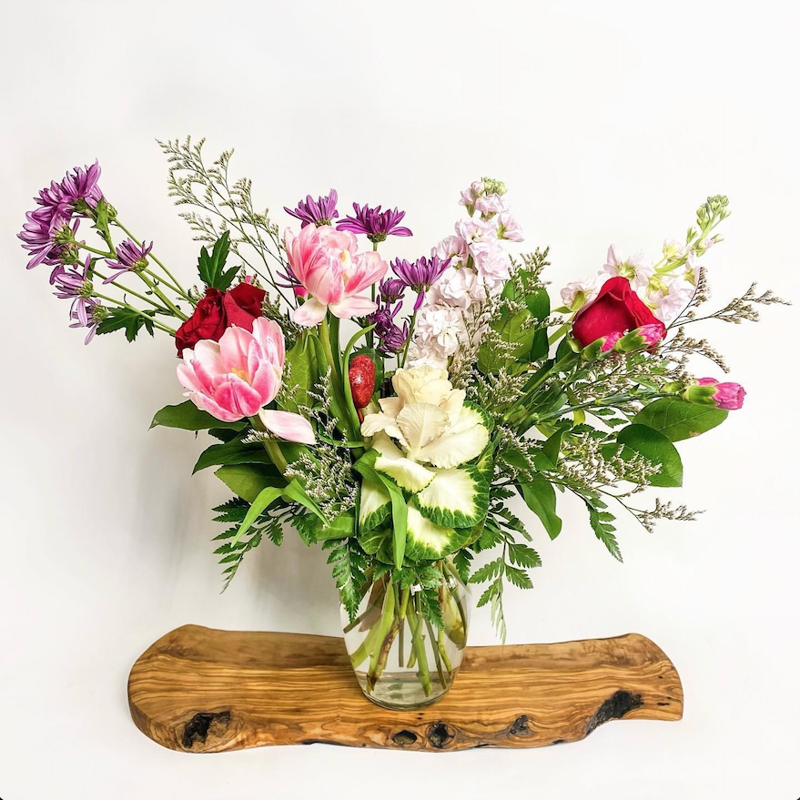 A glass vase filled with a colorful bouquet of fresh flowers including pink tulips, red roses, purple asters, white carnations, and green foliage, placed on a rustic wooden surface against a plain white background.