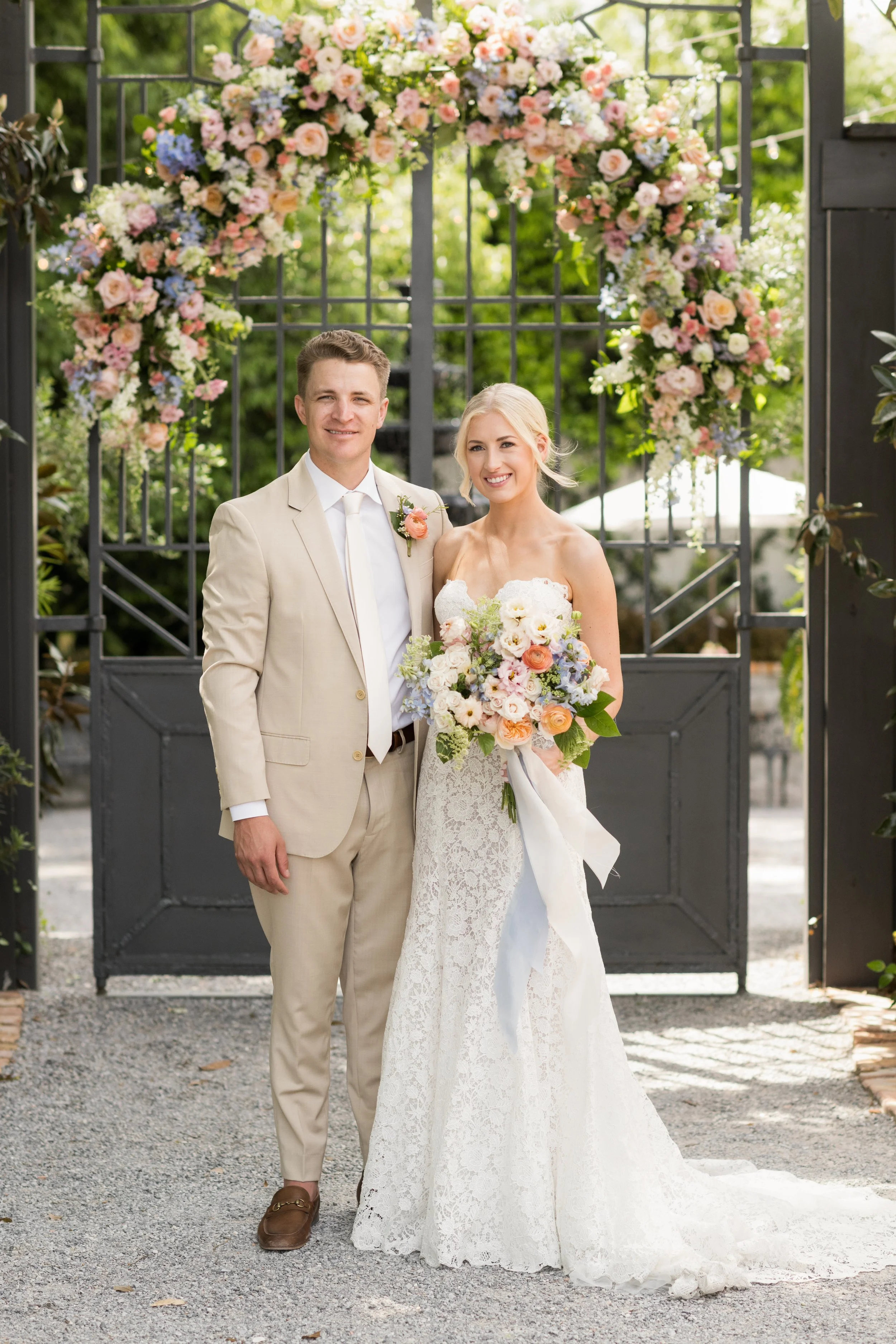 A bride and groom stand together outdoors on their wedding day in front of a floral arch and black gate, smiling for the camera. The bride has blonde hair and wears a lace wedding dress, holding a bouquet of pastel-colored flowers. The groom has shor