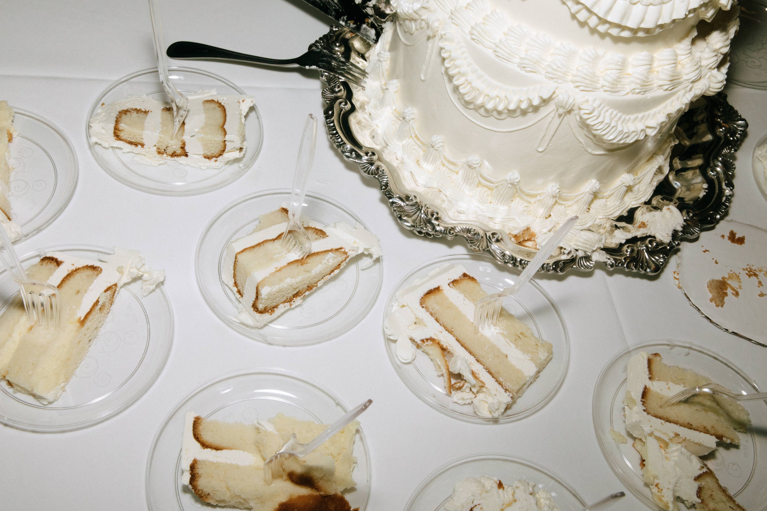 A table with slices of vanilla cake on clear plates, with forks, and a large decorated white cake with piped icing on a silver tray.