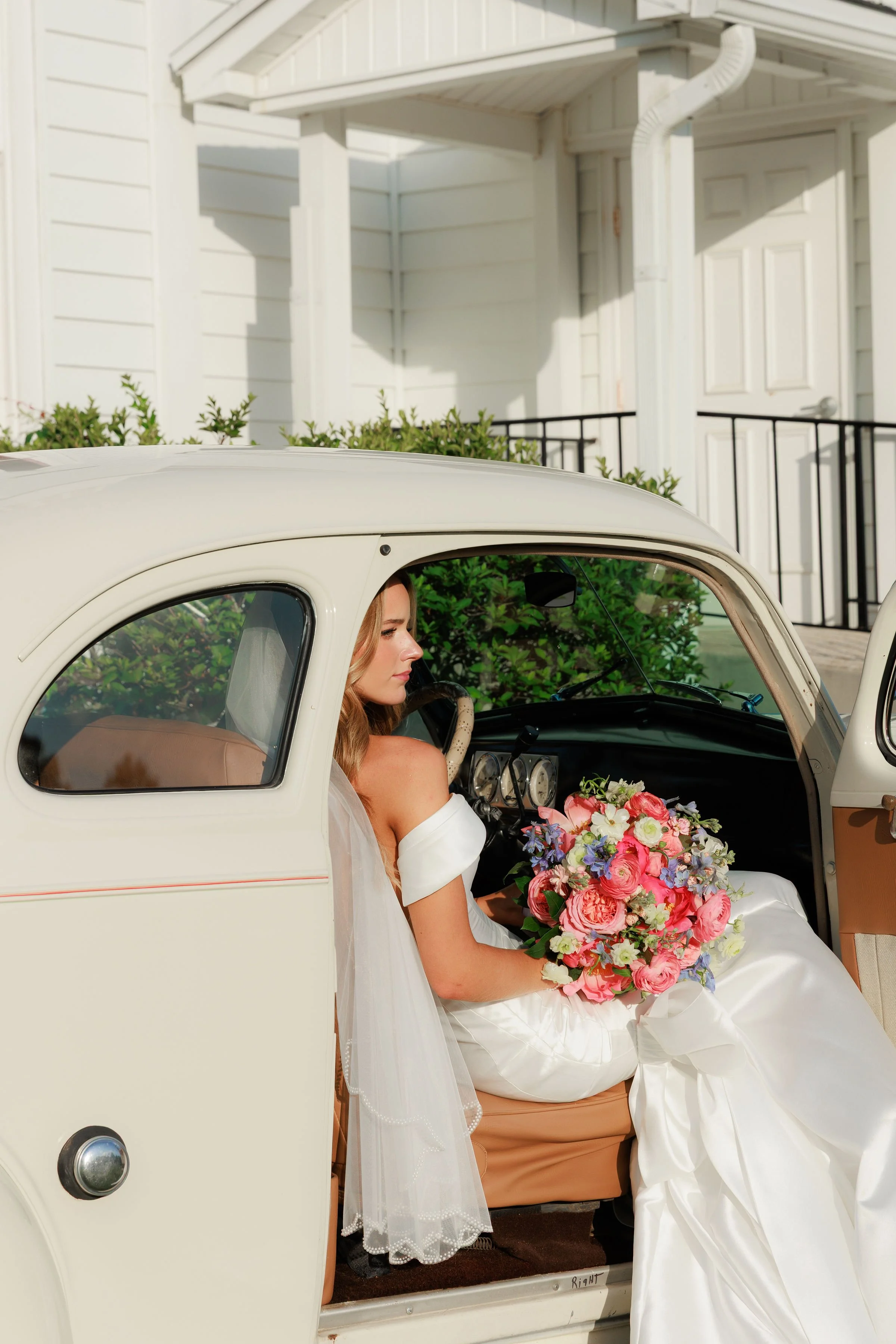 A bride sitting in a vintage white car holding a bouquet of pink, purple, and white flowers.