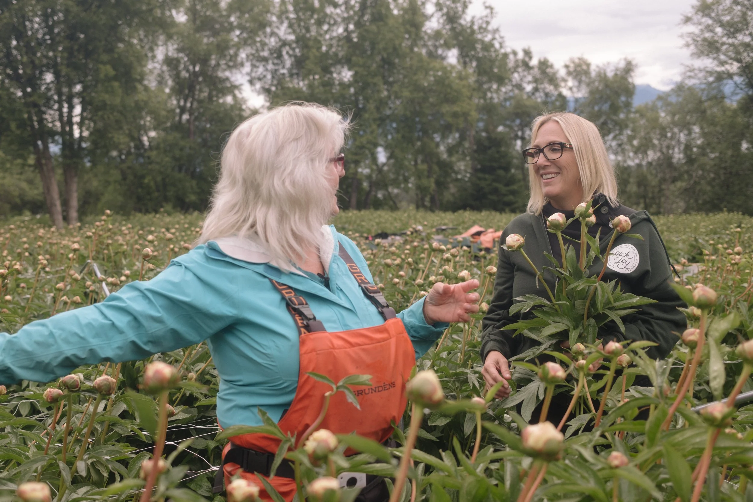 Two women talking and smiling in a flower field with trees in the background.