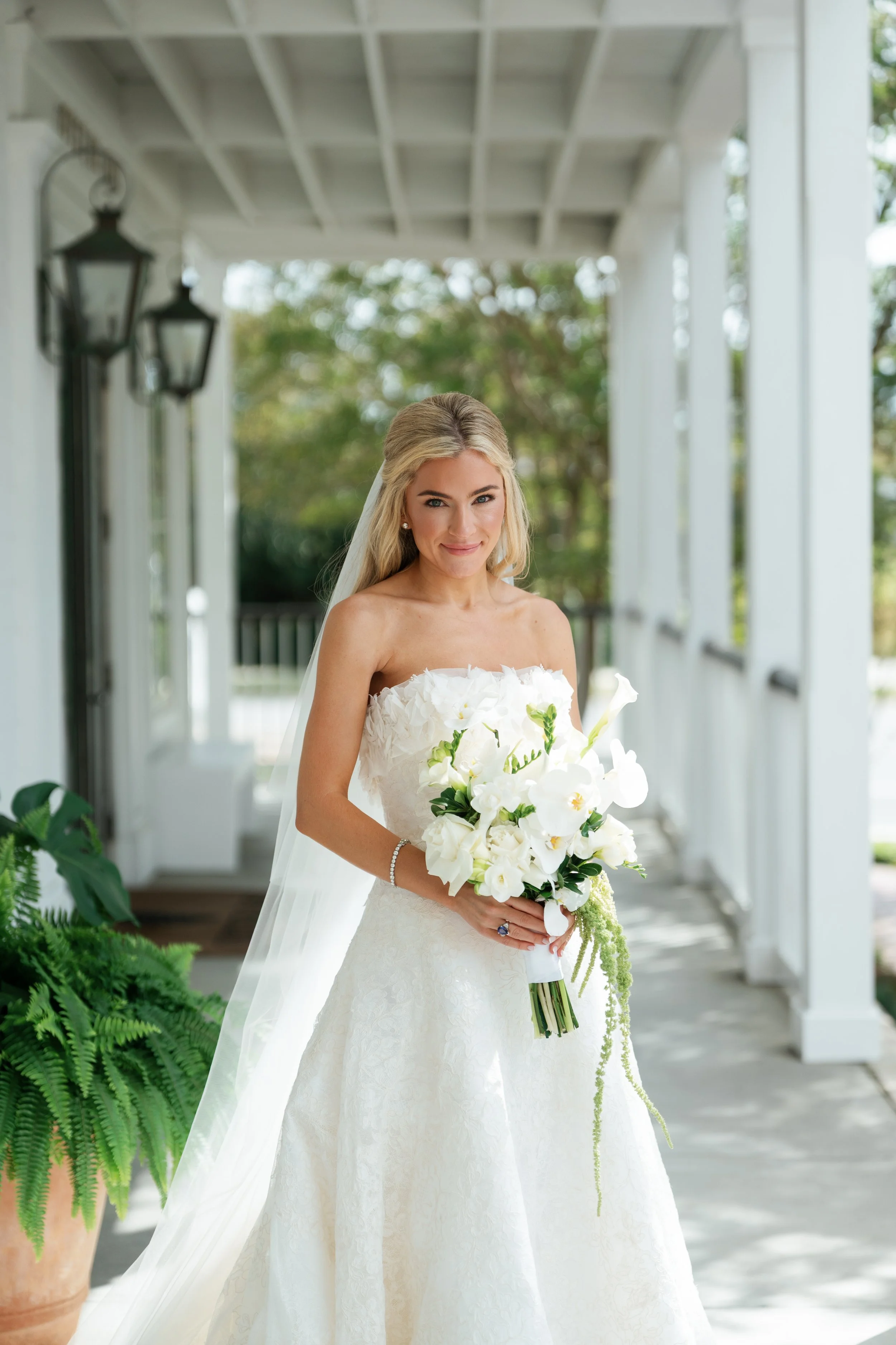 A bride in a white wedding dress holds a large bouquet of white flowers, standing on a porch with greenery in the background.