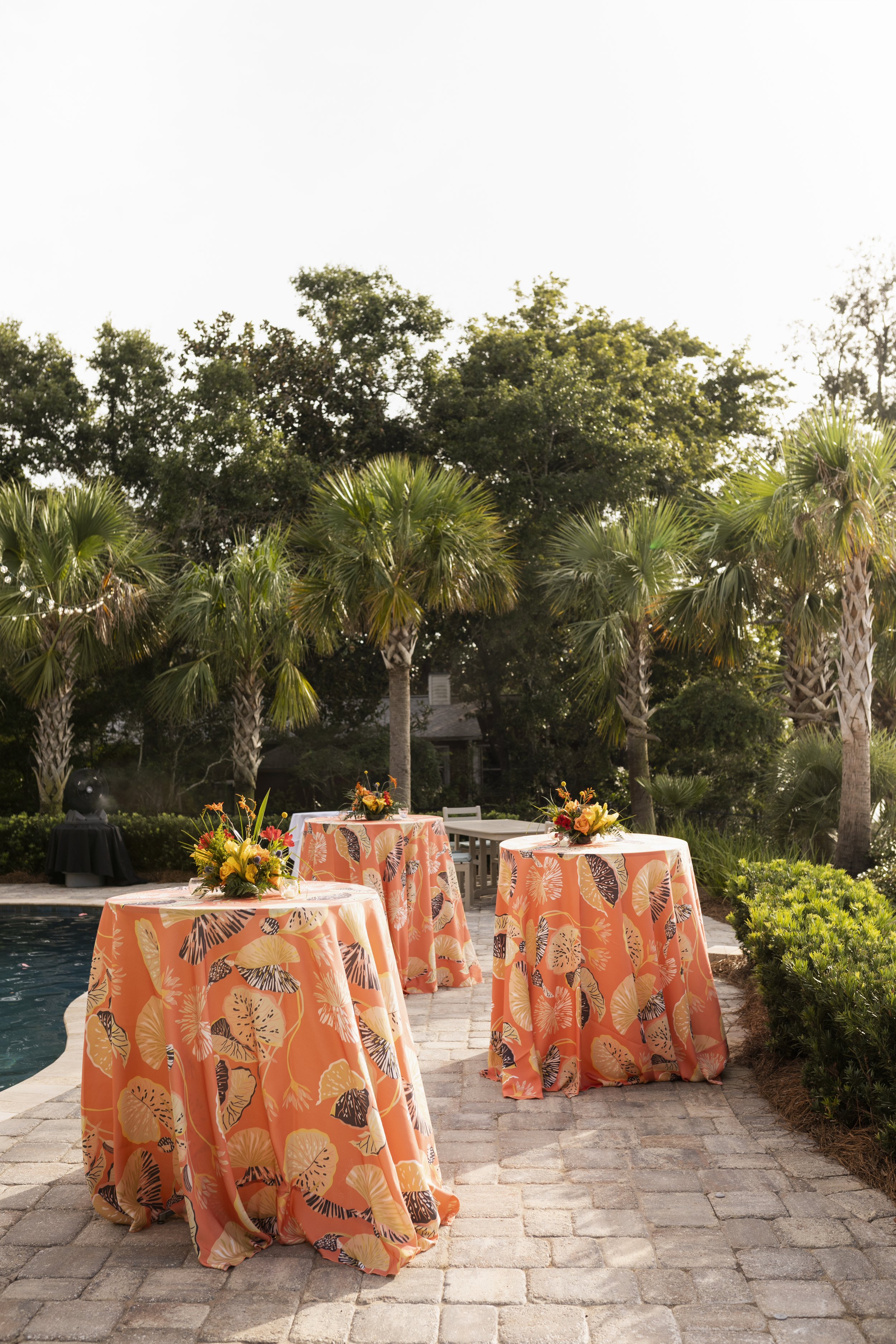 Three high tables with orange tablecloths adorned with leaf patterns, decorated with colorful floral arrangements, are set outdoors near a swimming pool surrounded by lush greenery and palm trees.