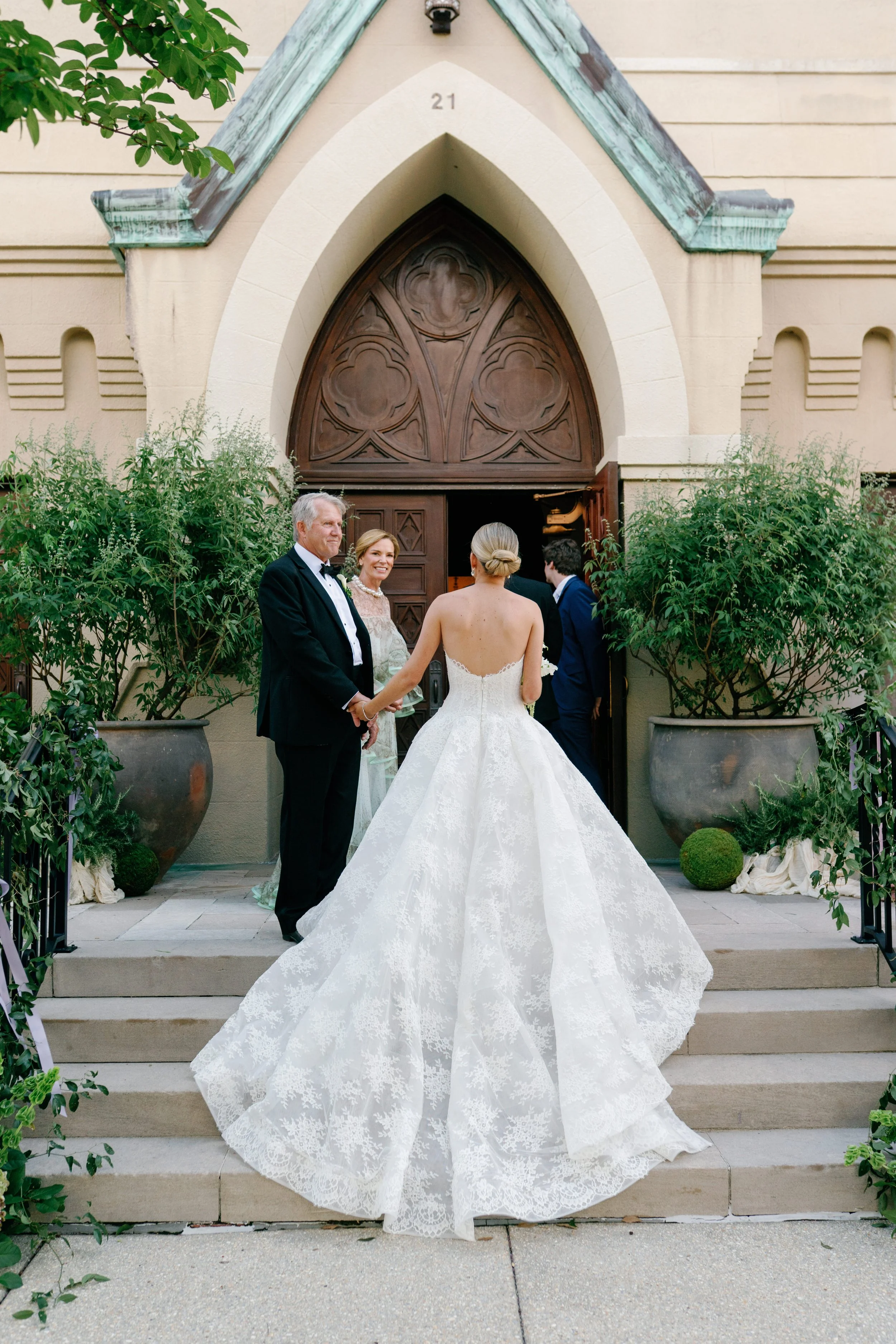 A bride in a white wedding gown stands on steps outside a church, holding hands with an older man, possibly her father, as she prepares to walk down the aisle. Two other guests, a woman and a man, are also present near the open church door, which has