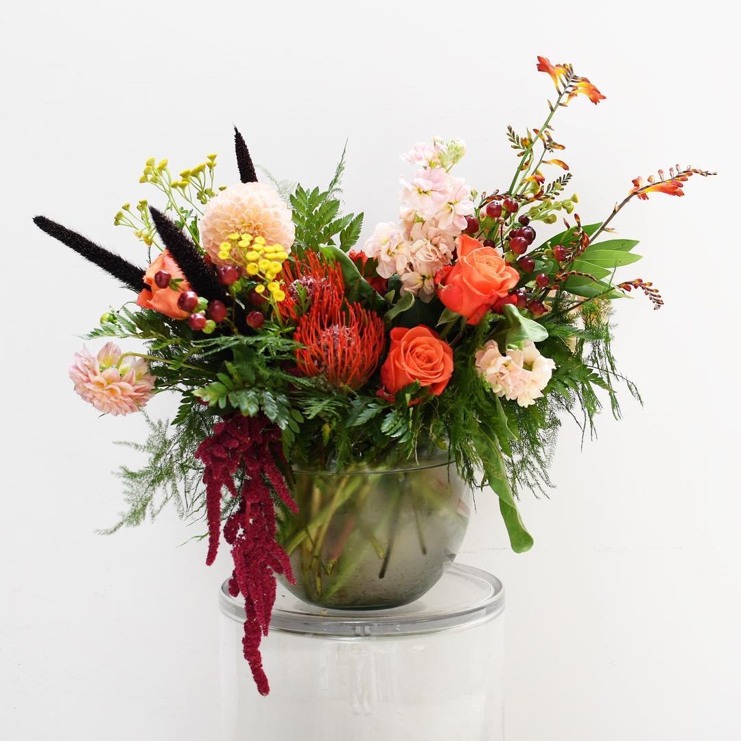 A colorful floral arrangement in a glass vase with pink, orange, and red flowers, set against a plain white background.