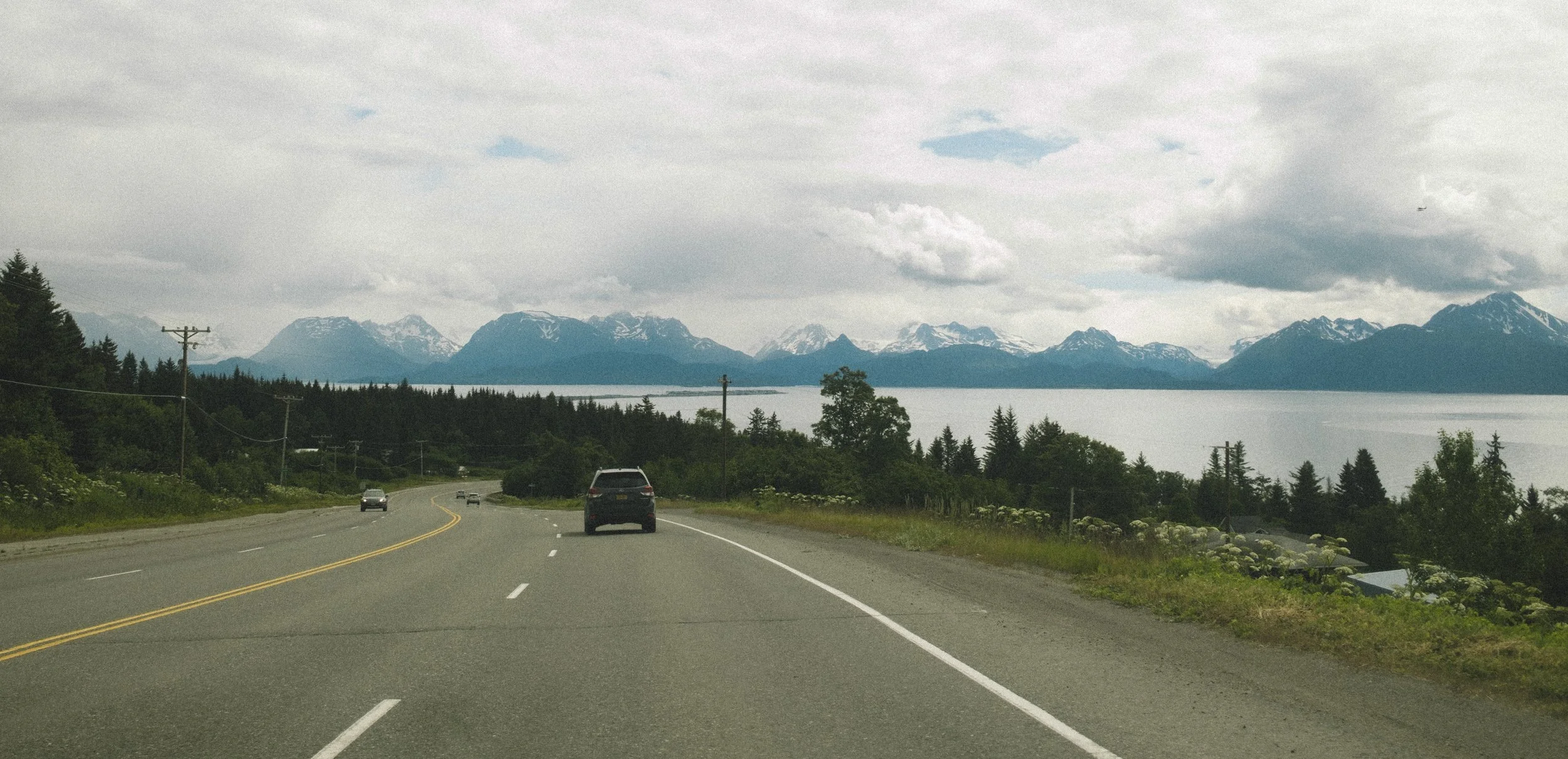 Scenic view of a winding two-lane road with a car driving, surrounded by trees and wildflowers, overlooking a large lake with snow-capped mountains in the distance under cloudy sky.