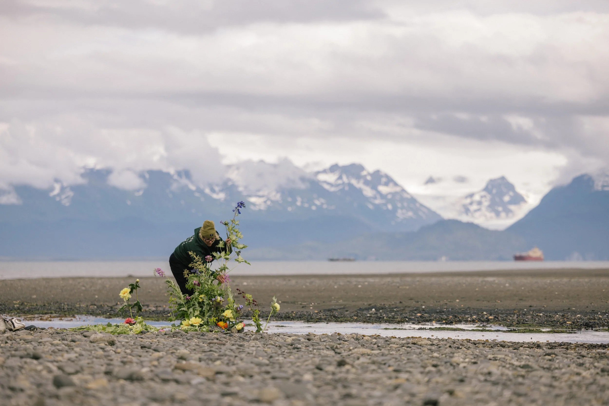 A person placing flowers on a memorial or grave on a rocky beach with snow-capped mountains and a cloudy sky in the background.