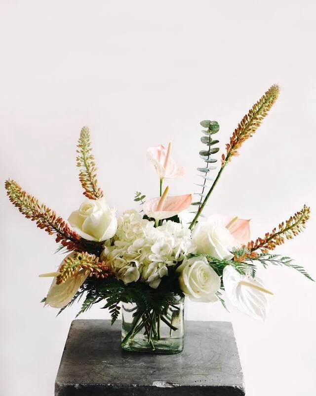 A floral arrangement in a clear glass vase with white roses, hydrangeas, and pink anthuriums, accented with green eucalyptus and tall, spiky grass, placed on a dark gray pedestal against a white background.