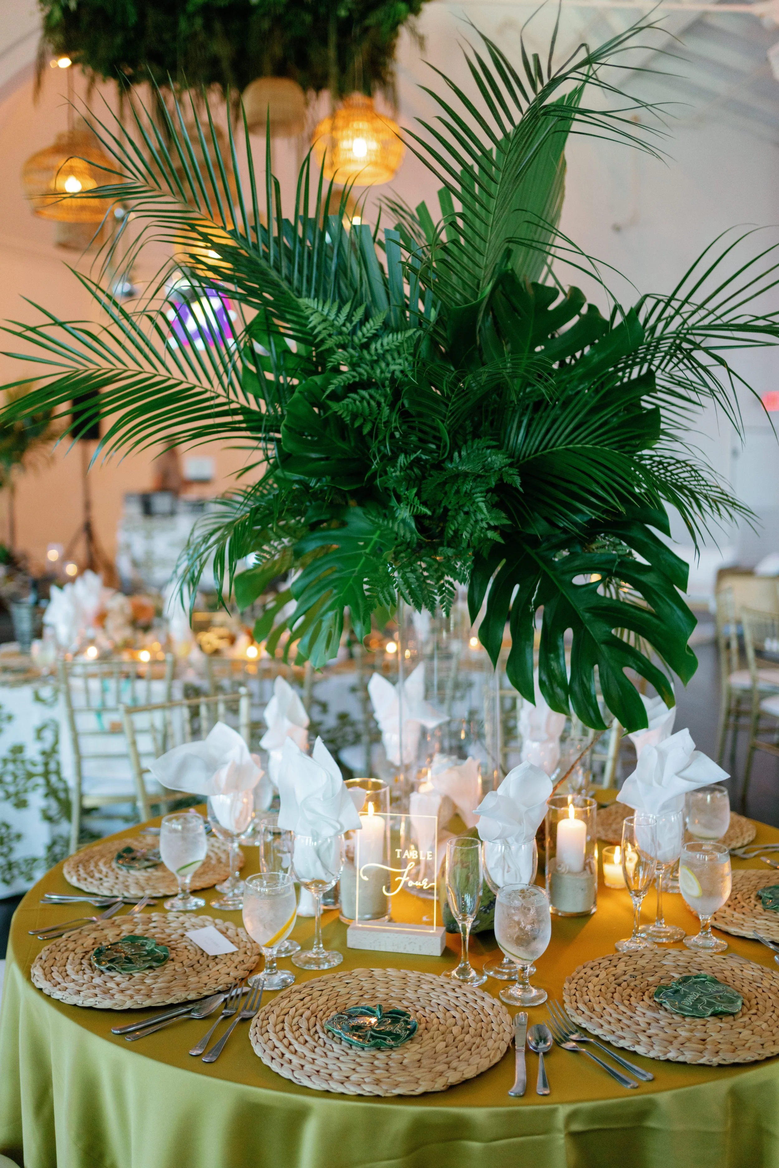 A decorated banquet table with a tall lush green tropical plant centerpiece, woven placemats, clear glassware, white napkins, candles, and a table sign that reads 'Table Four' in gold lettering. The setting suggests a festive or elegant event.