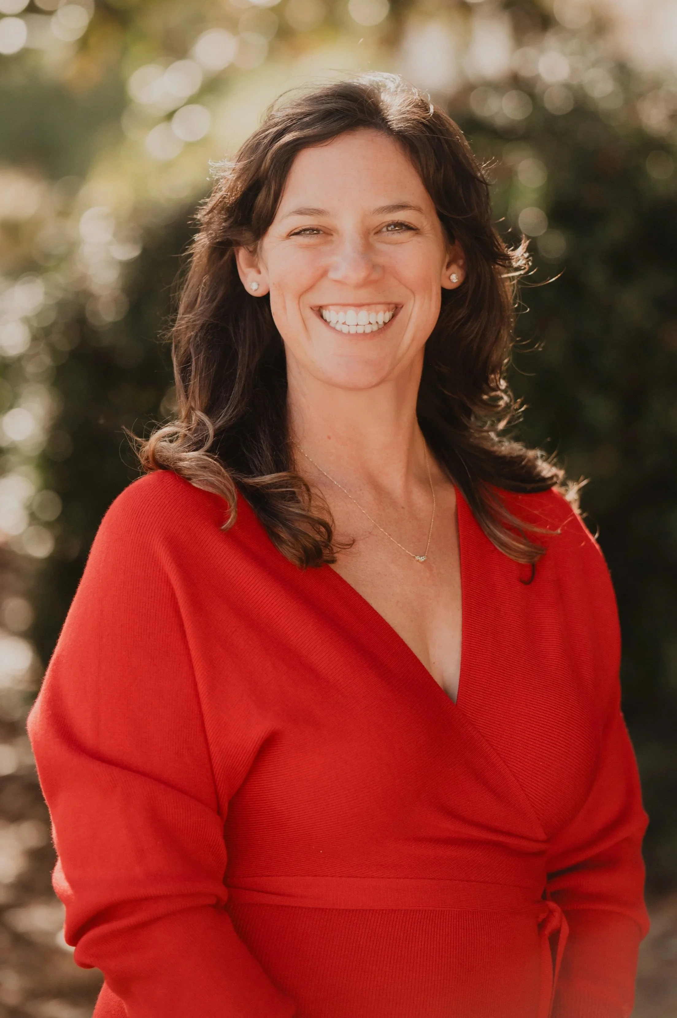 A woman with dark brown, wavy hair smiling outdoors, wearing a red dress and small earrings.