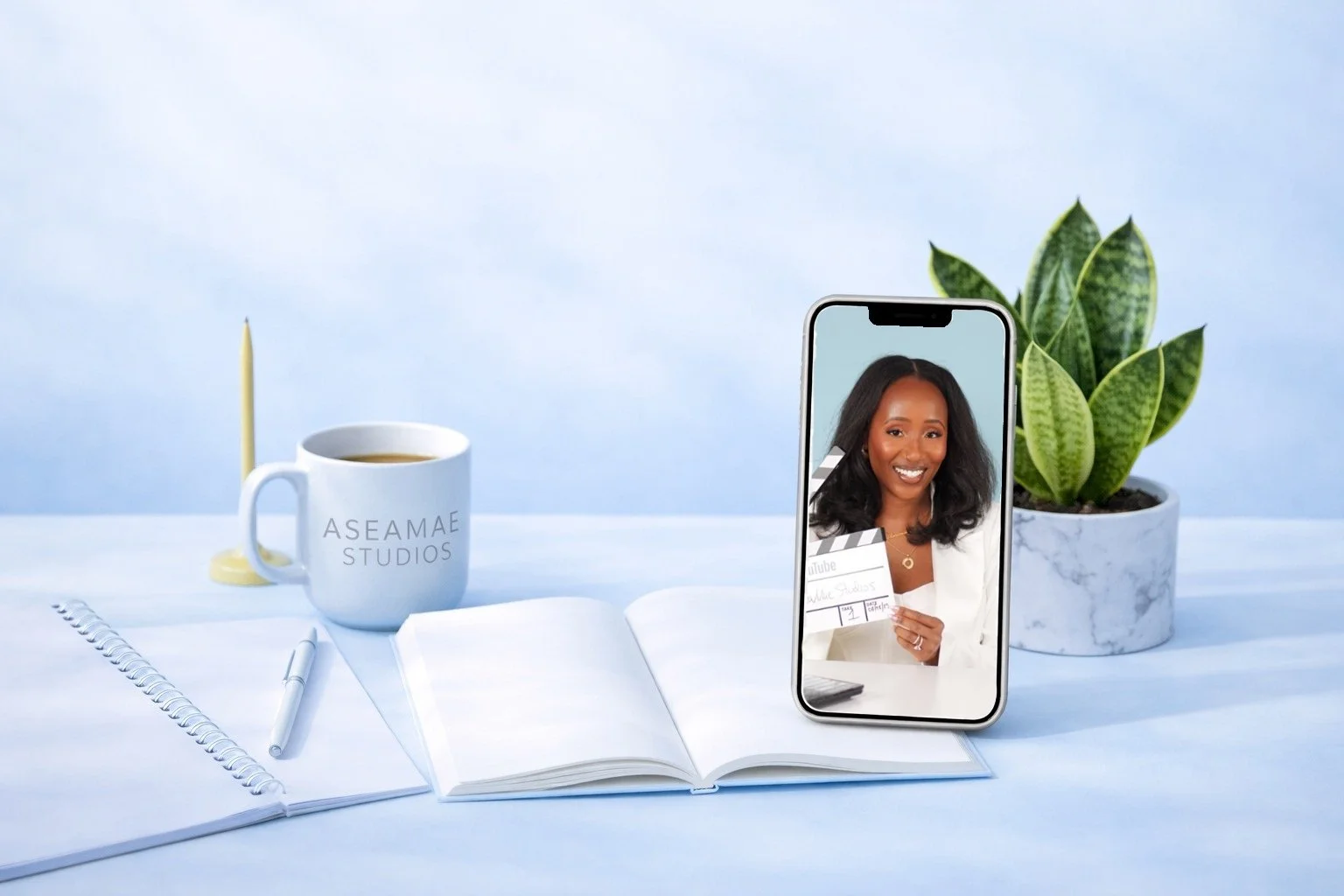 A workspace setup with a white notebook, pen, and a mug labeled 'ASEAMAE STUDIOS' on a white surface. A potted plant with green leaves is on the right. A smartphone displays a woman smiling and holding a clapperboard, with a blue sky background.