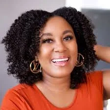 A woman with curly black hair wearing gold hoop earrings and an orange top, smiling at the camera.