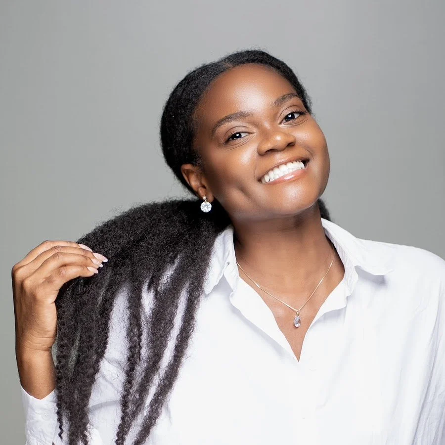 A smiling woman with dark skin, black textured hair, dressed in a white shirt with jewelry, against a gray background.