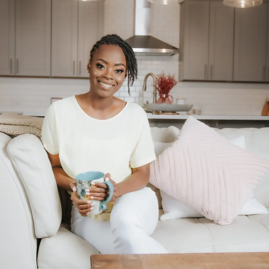 A woman sitting on a cream-colored couch in a modern kitchen, holding a mug, smiling at the camera.