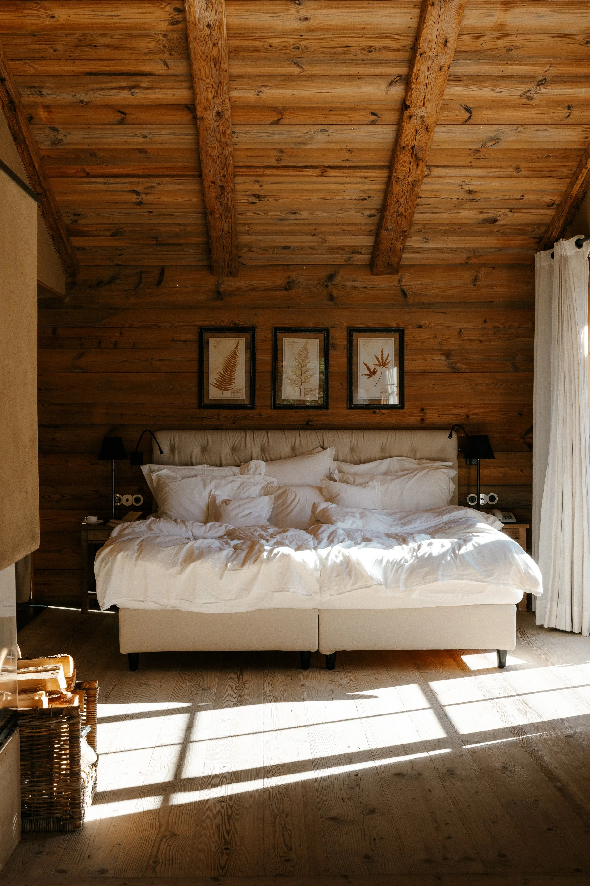 A cozy bedroom with a large, unmade bed, white bedding, and multiple pillows. The walls and ceiling are made of wood, with rustic beams on the ceiling. There are three framed leaf prints hanging above the bed. Sunlight streams in through white curtains on a window to the right, casting shadows on the wooden floor.