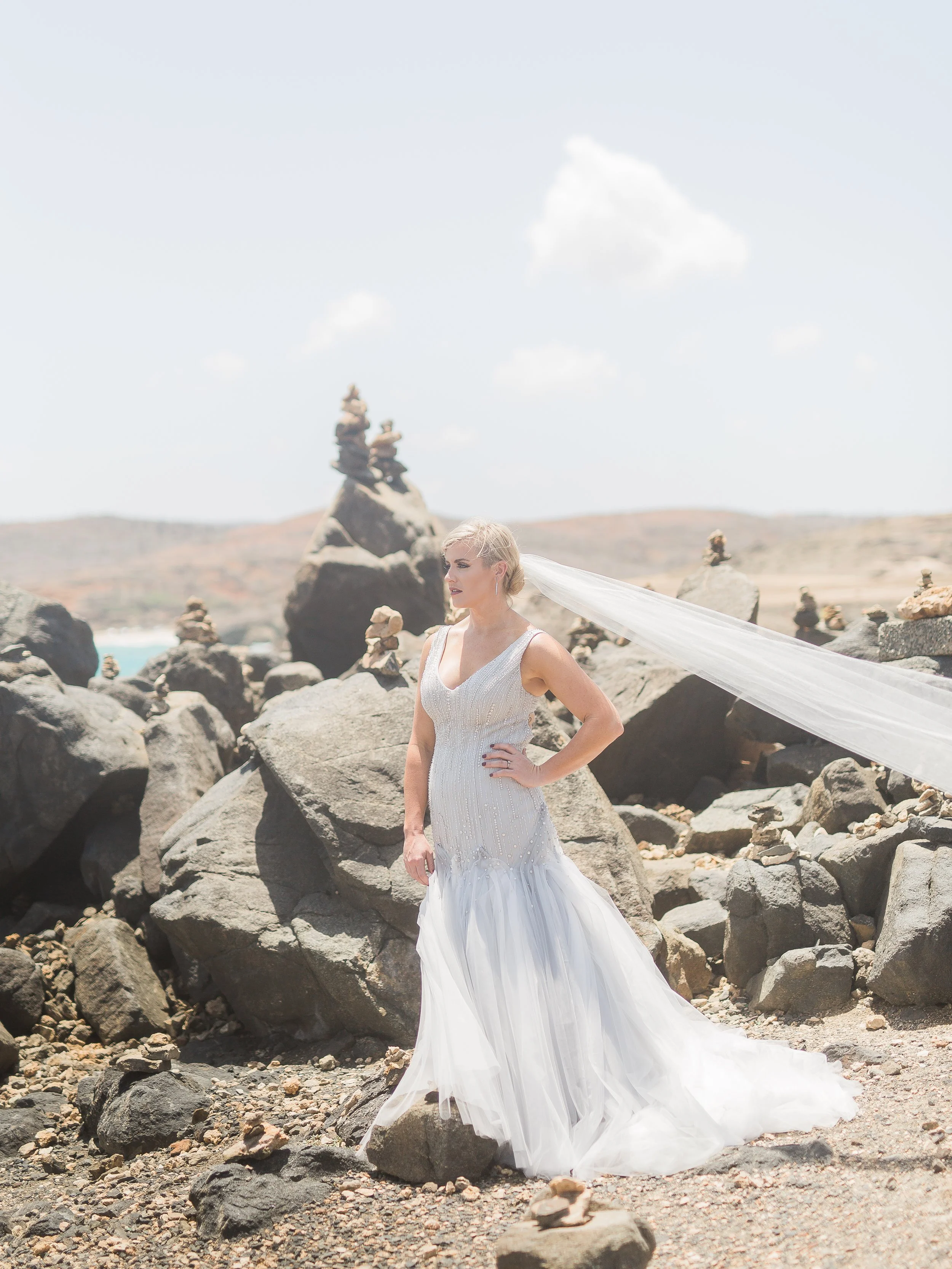Bride stands on the north shore of Aruba, surrounded by rock formations in a custom wedding dress by Linh's bridal with cathedral veil blowing in the Aruba wind. Photo by Danielle Elizabeth Weddings http://www.danielleelizabethweddings.com